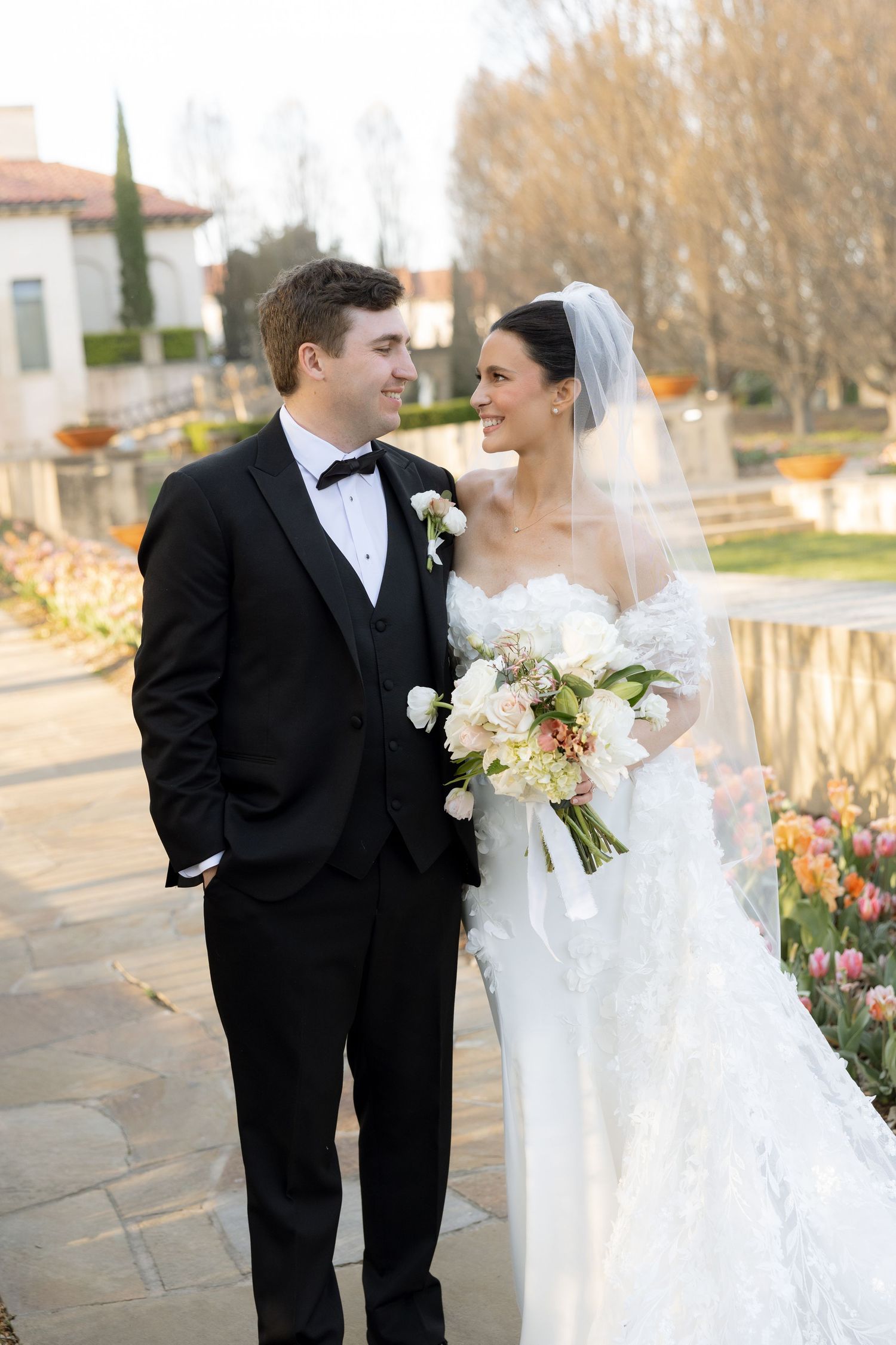 Smiling bride in lace gown with veil and bouquet stands with groom in tuxedo at garden venue.