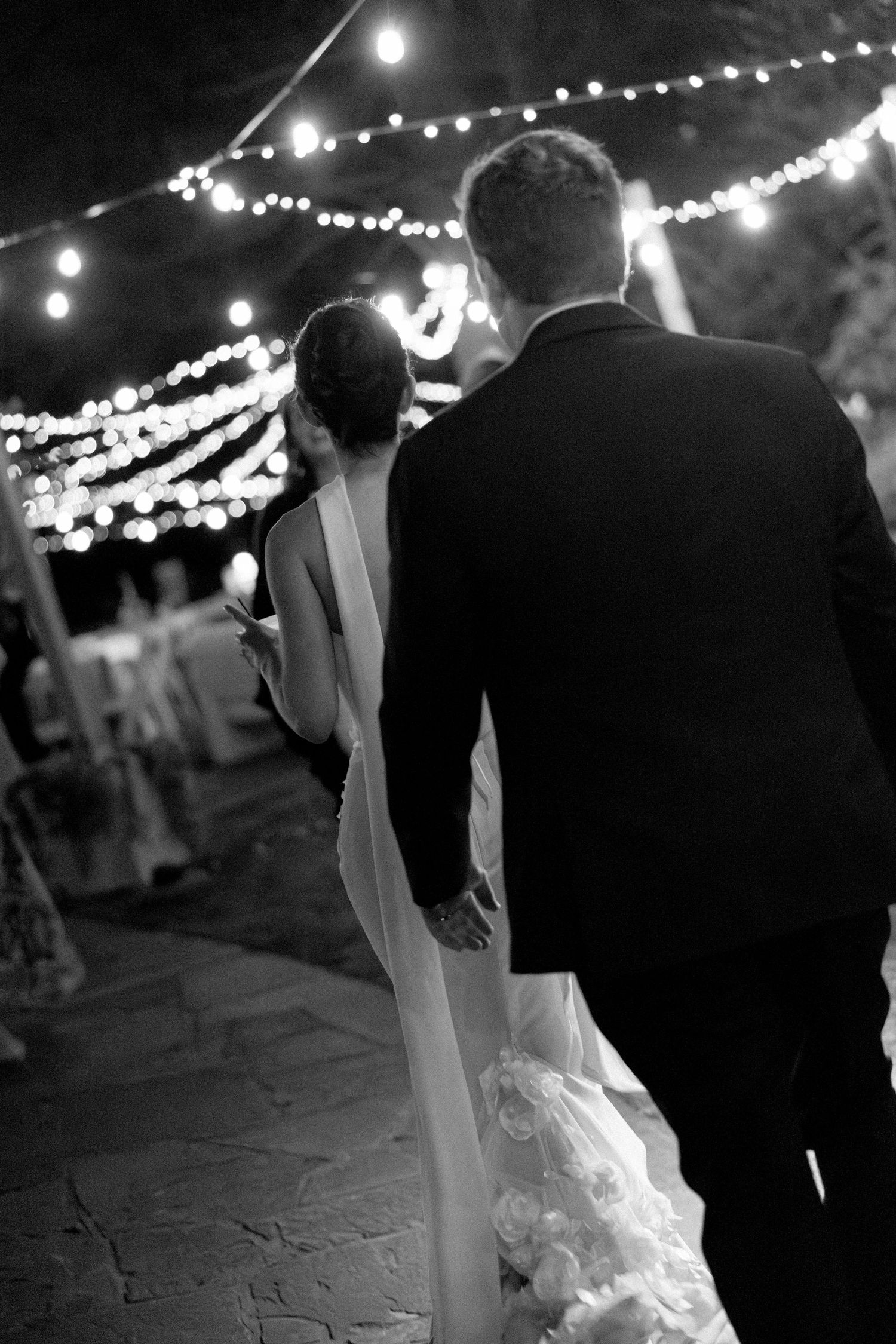 Black and white photo of bride and groom walking under string lights at outdoor wedding reception.