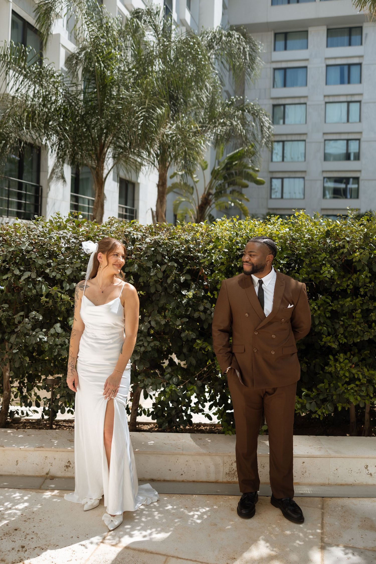 Wedding couple in elegant attire pose against tropical landscaping and modern architecture.