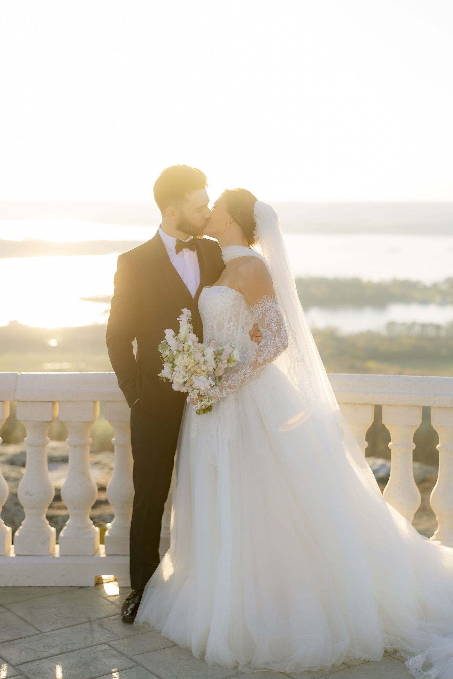 Bride and groom share a romantic kiss on a sunlit balcony, with a stunning waterfront view in the background.