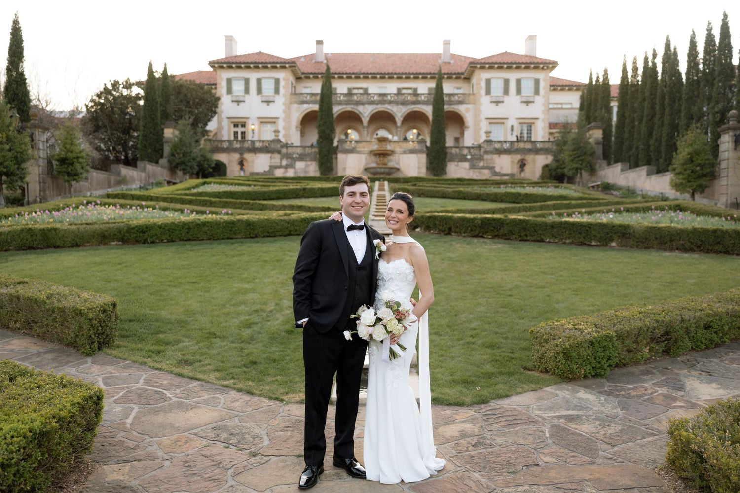Bride and groom posing together in front of elegant Italian-style mansion with manicured gardens.
