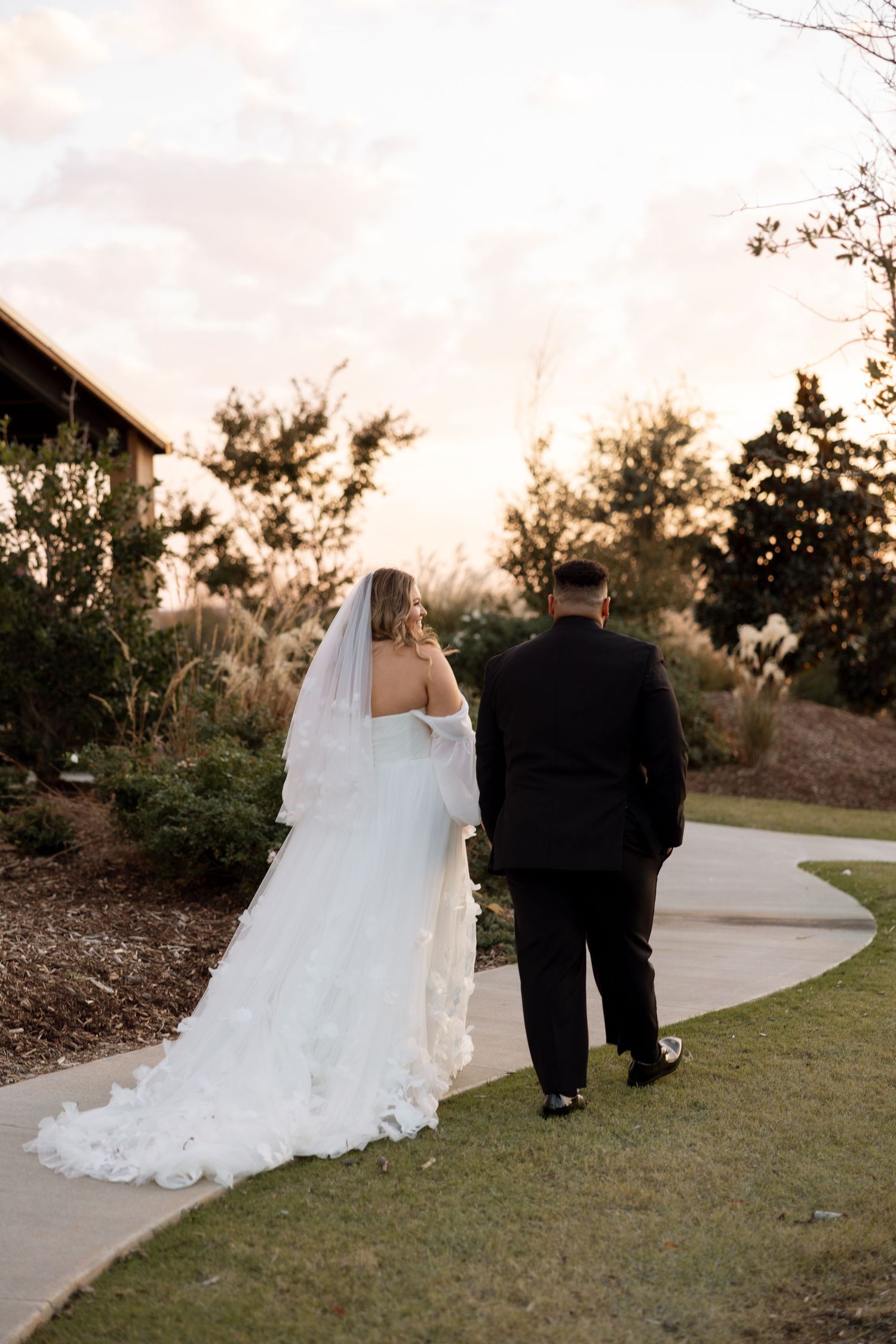 Couple walks along garden path at sunset with flowing wedding dress train.