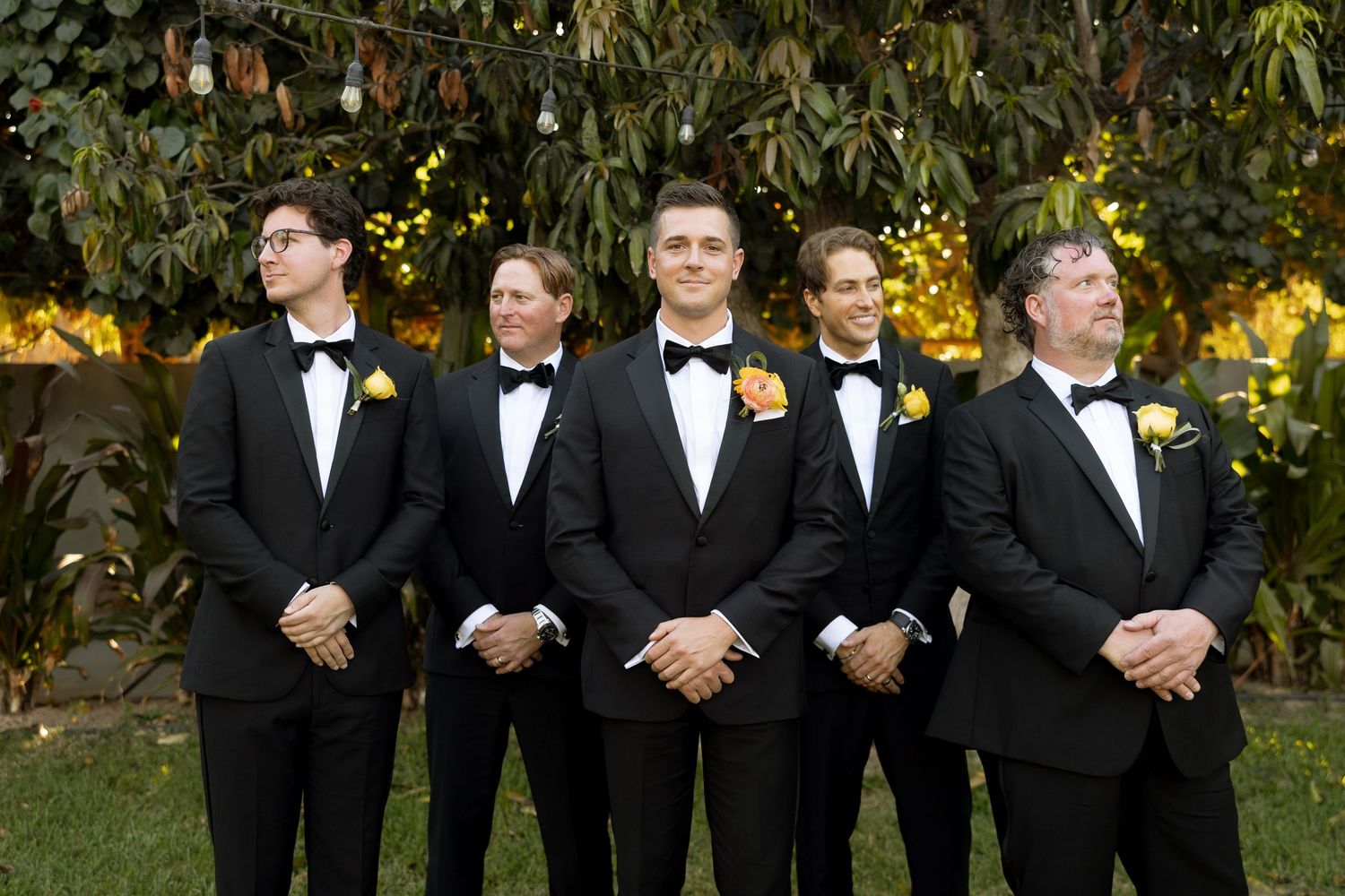 Five groomsmen in black tuxedos with yellow boutonnieres pose outdoors amid lush greenery at a wedding.