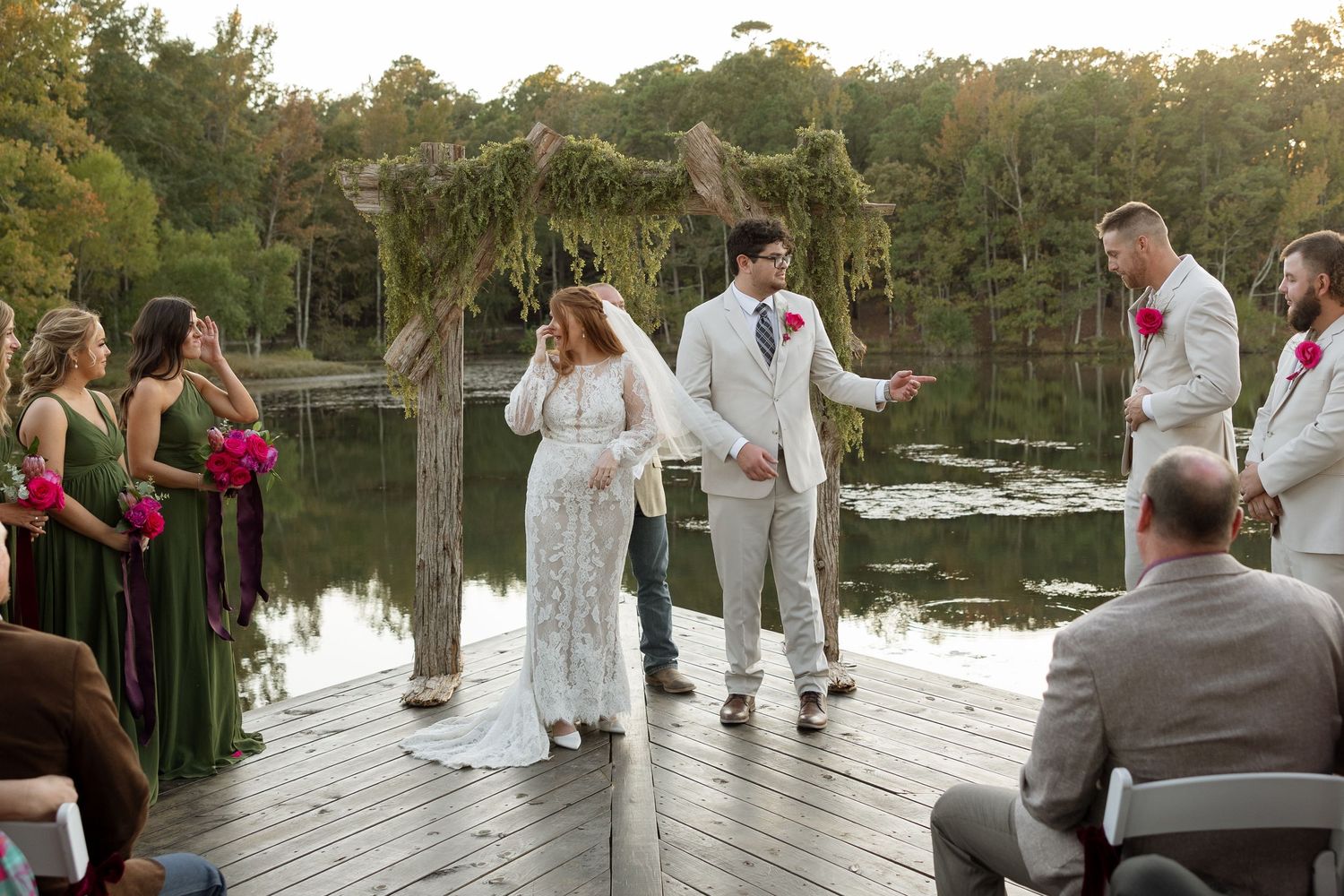 A wedding ceremony takes place on a lakeside dock with a rustic arch and attendants in dark dresses.