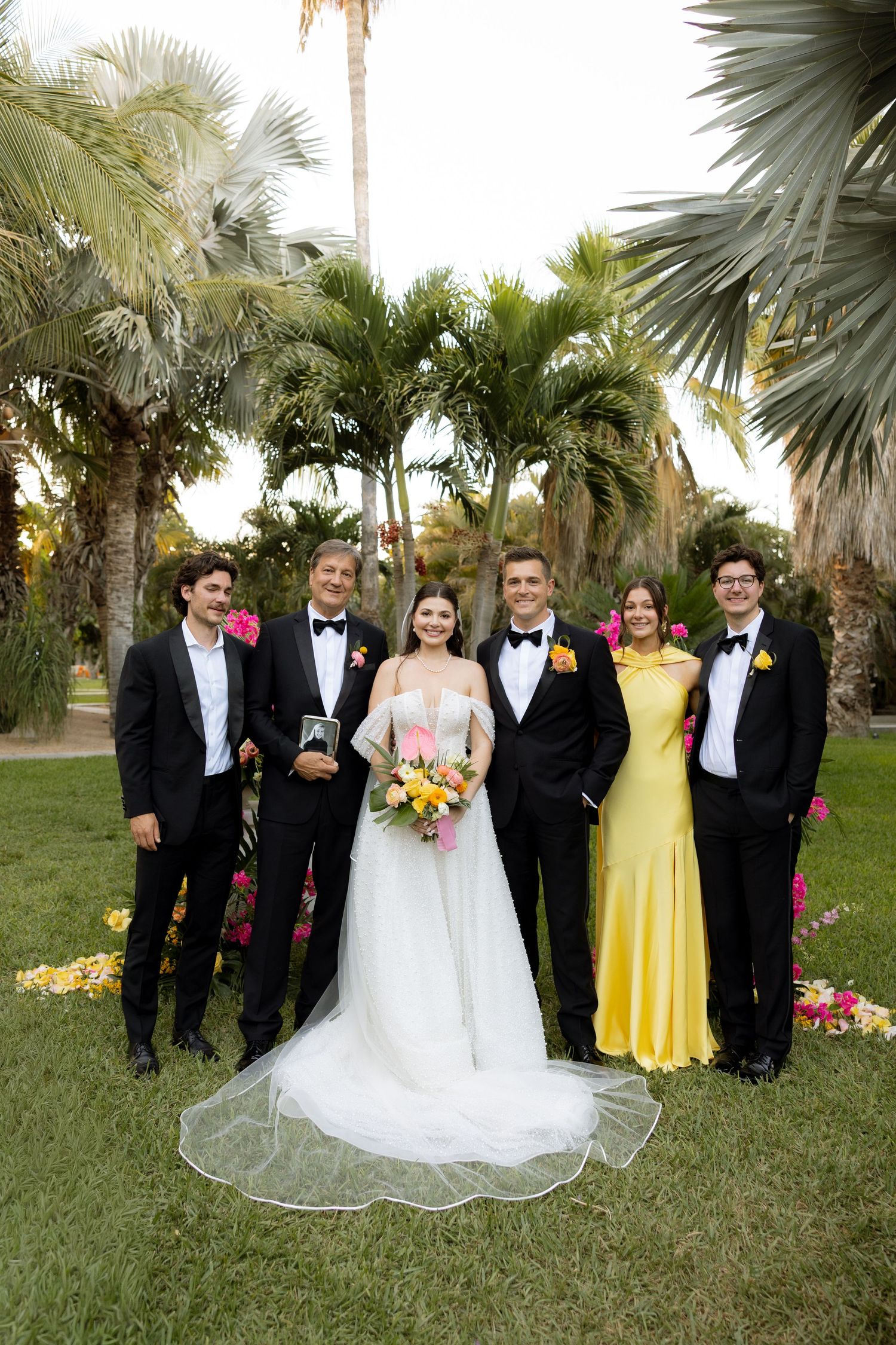 Wedding party poses outdoors with bride in white gown and groom, flanked by guests in tuxedos and yellow dress.