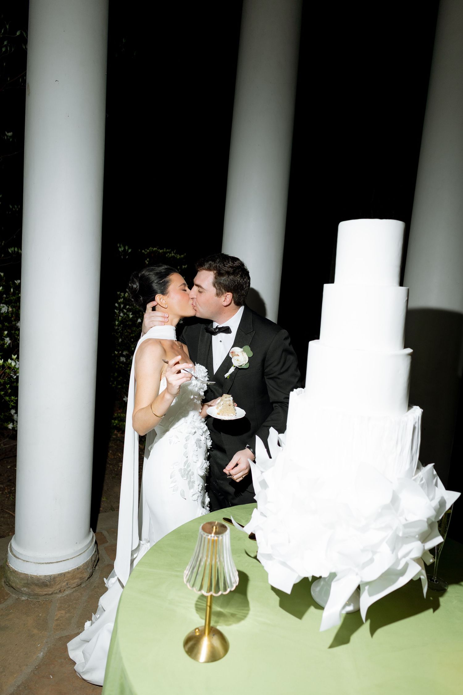 Bride and groom sharing a kiss beside tall white ruffled wedding cake between white columns at night.