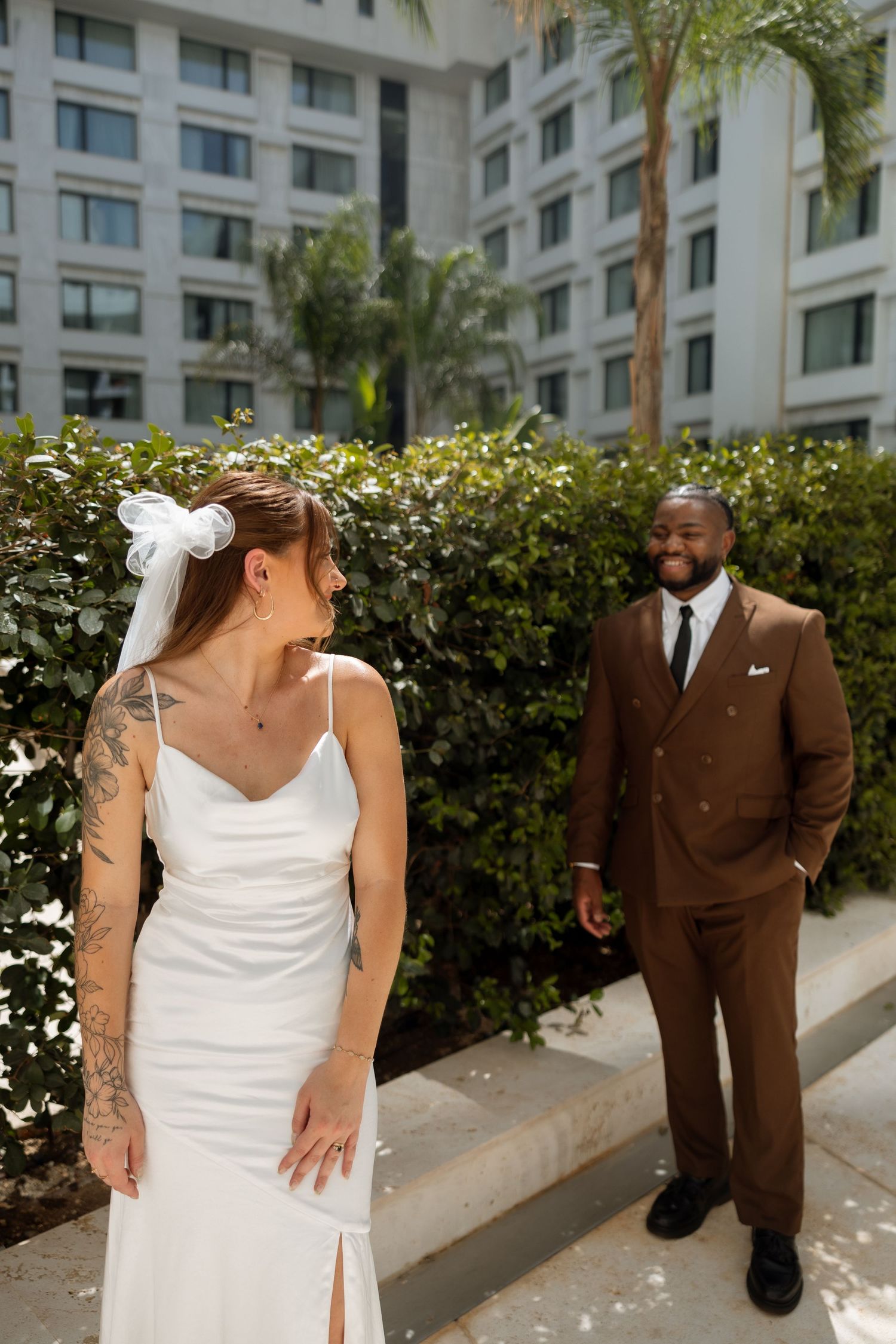 Bride in white dress with tattoos shares a joyful first look moment with groom in brown suit at hotel courtyard.