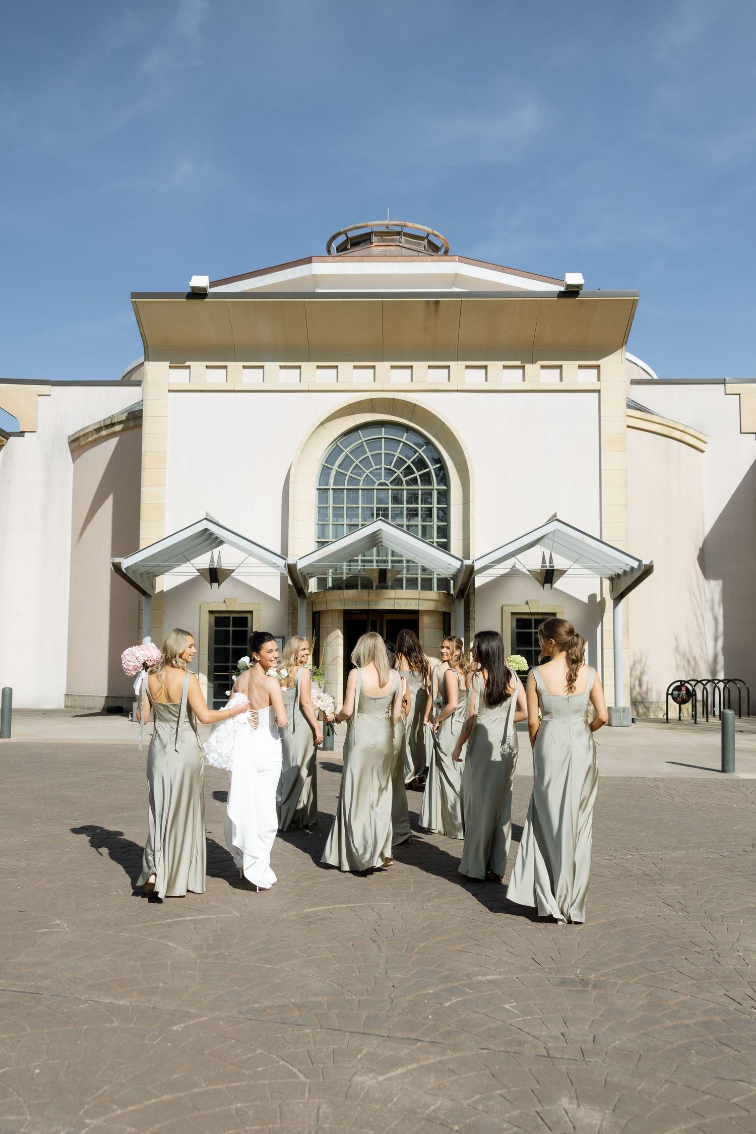 Bridesmaids in sage green gowns walk with bride toward elegant wedding venue entrance under blue sky.