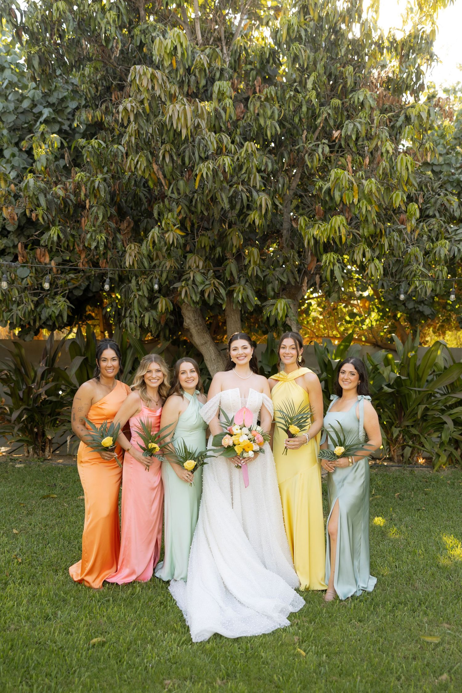 Bride in white gown poses with five bridesmaids in colorful mismatched dresses holding bouquets outdoors.