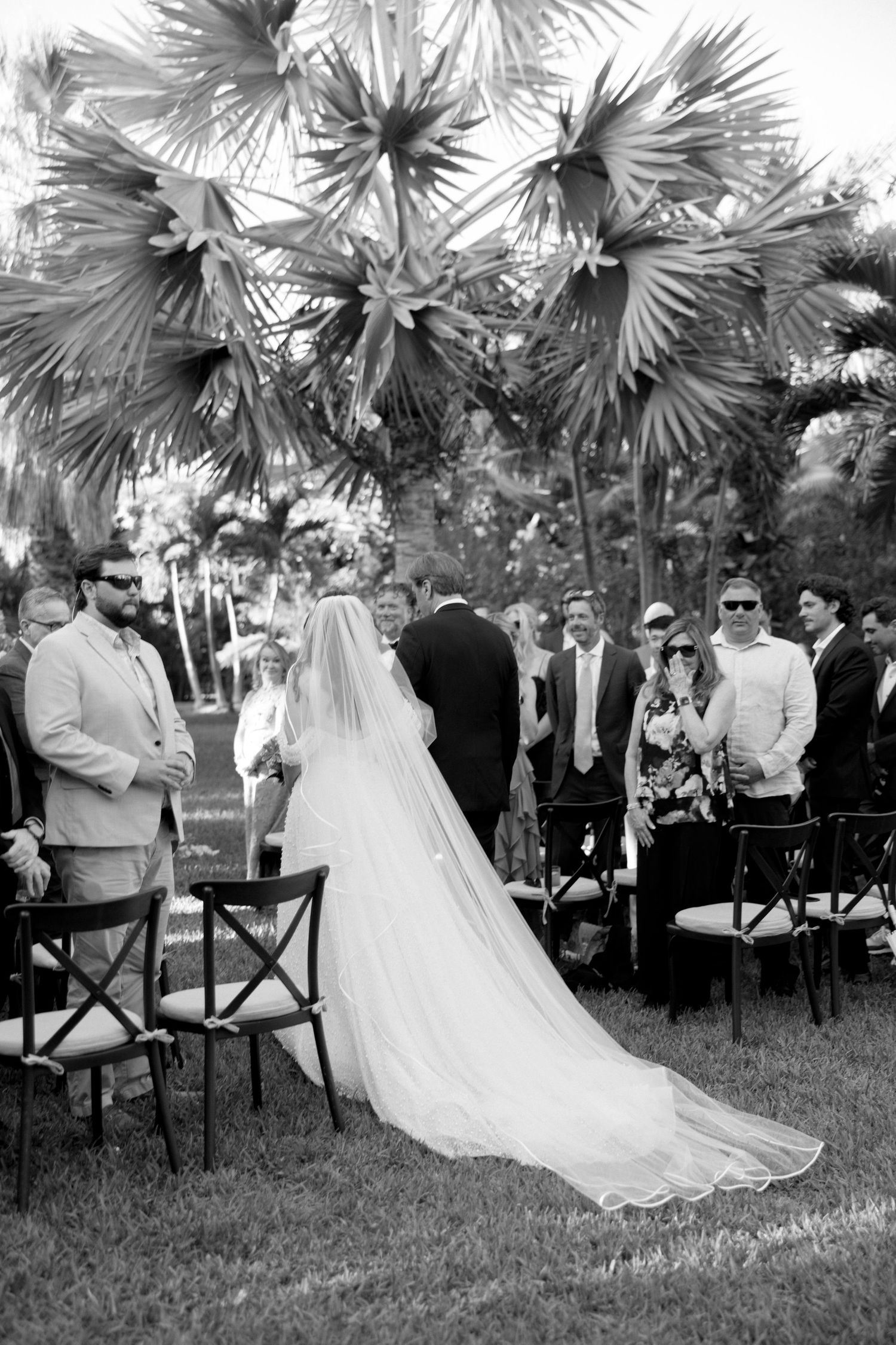 Black and white photo of bride in long veil walking down outdoor wedding aisle lined with guests.