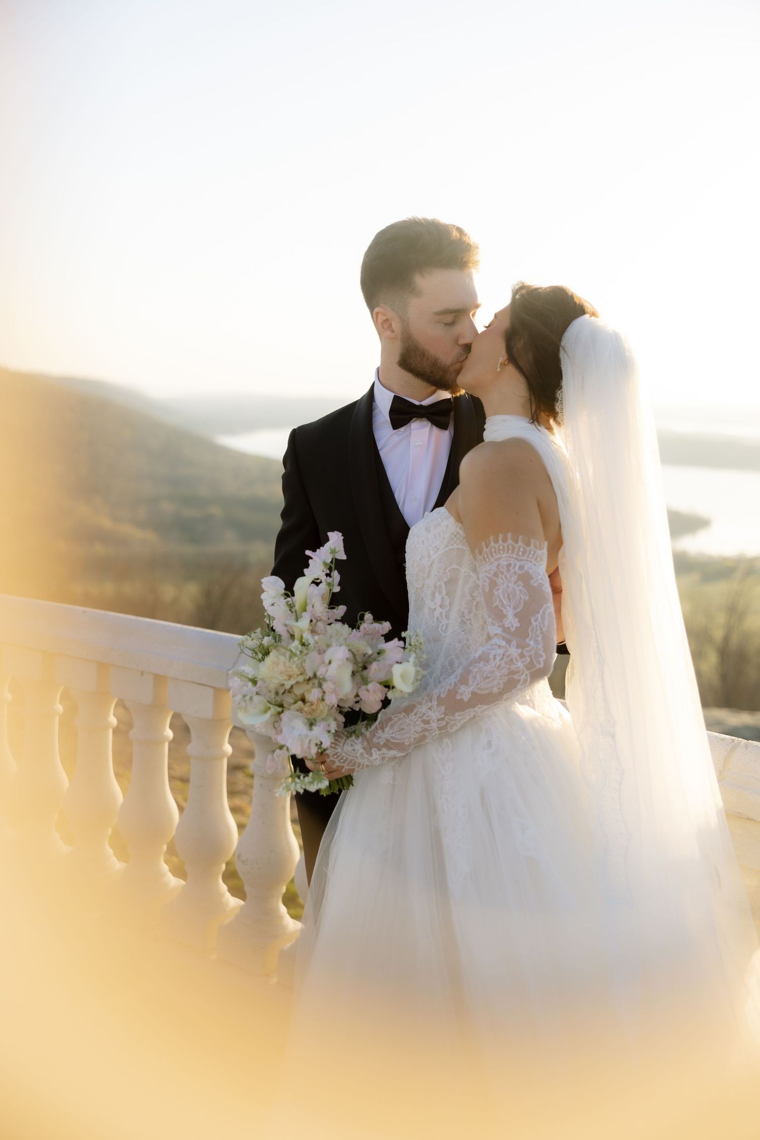 Bride and groom share a romantic kiss on a sunlit balcony, bride holding white floral bouquet with scenic views behind.