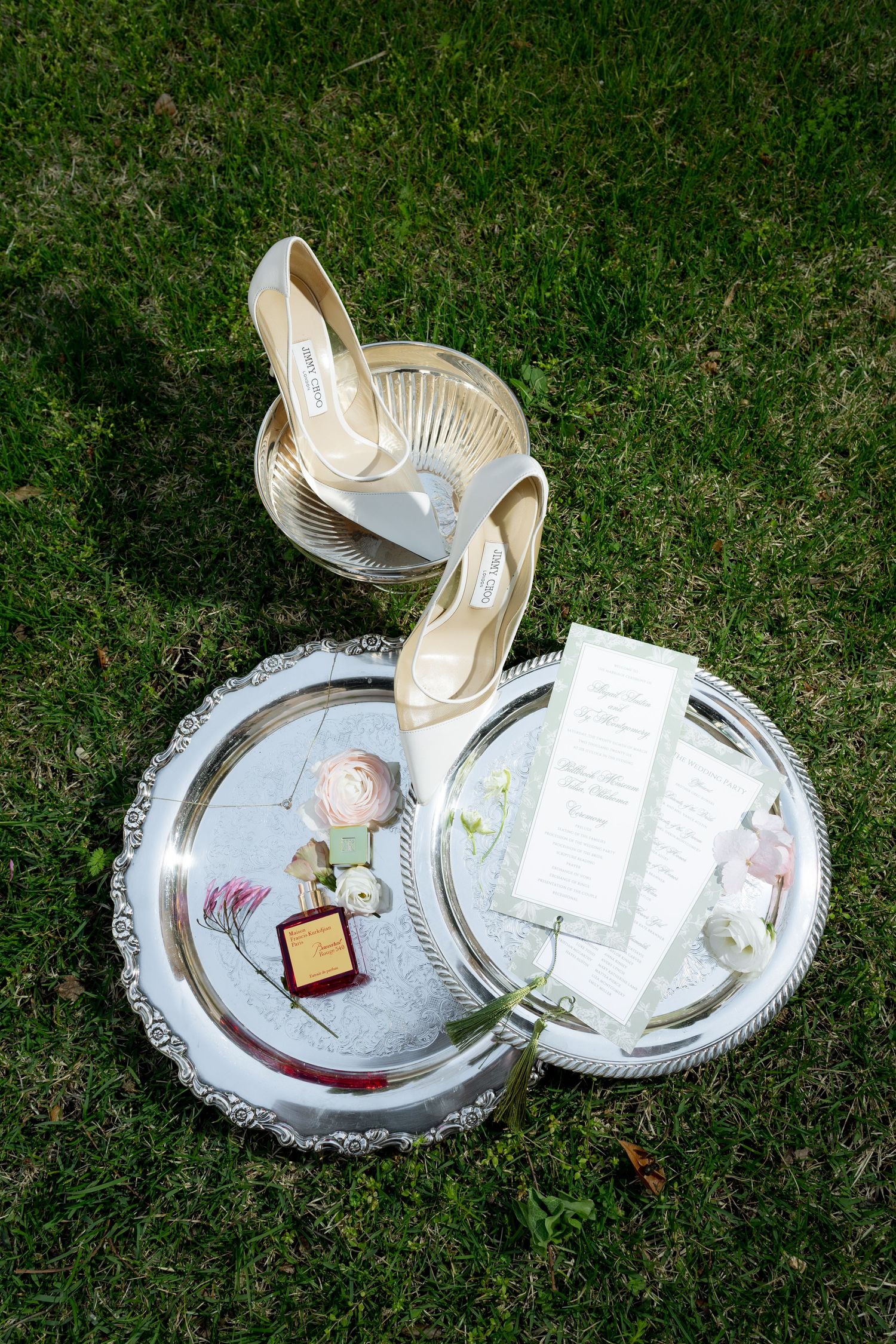 Wedding flat lay of Jimmy Choo heels, ring box, perfume, and program on silver tray on grass.