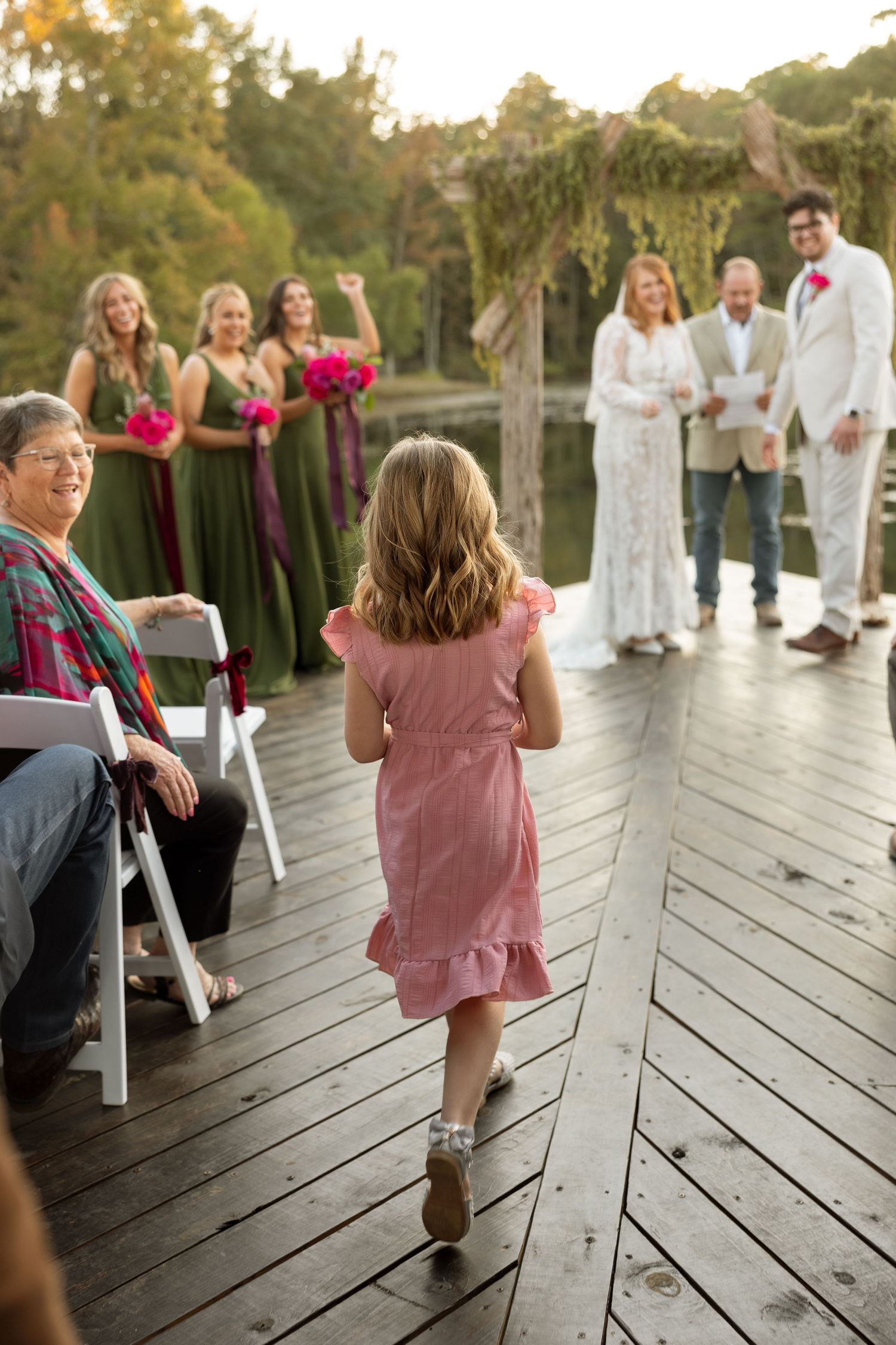 A young girl in a pink dress walks down the aisle during an outdoor lakeside wedding.