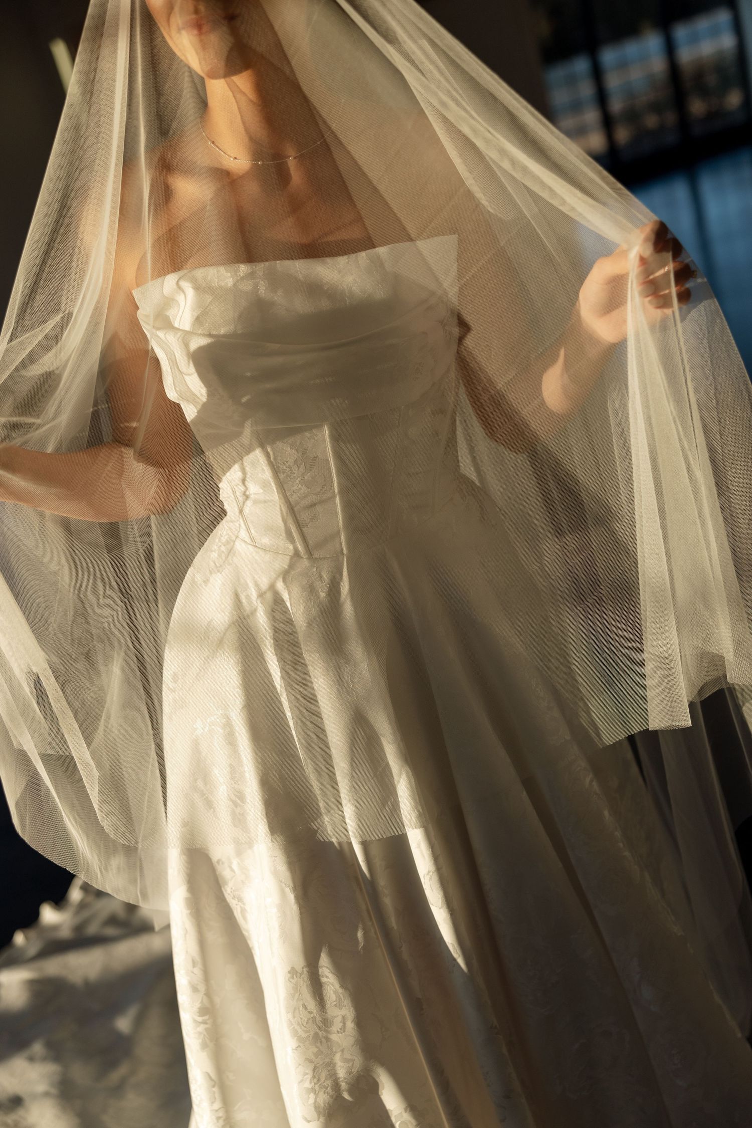 A bride in a classic white strapless wedding gown holds a delicate tulle veil in soft natural lighting.