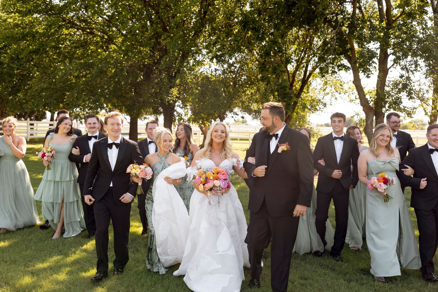 A wedding party celebrating outdoors with bridesmaids in sage green dresses and groomsmen in black tuxedos.