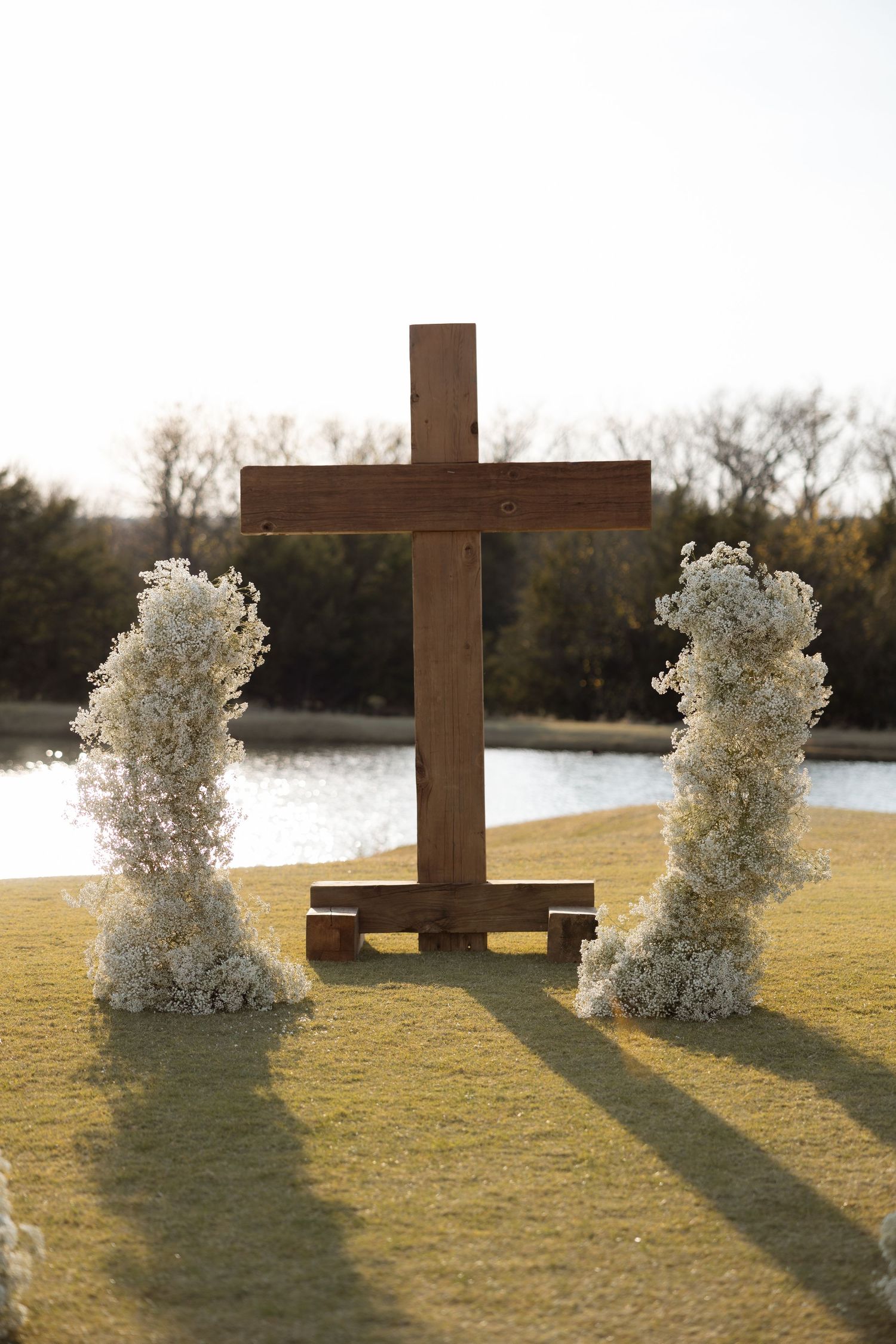 Large wooden cross with white floral arrangements on a grassy lawn near a lake at sunset.