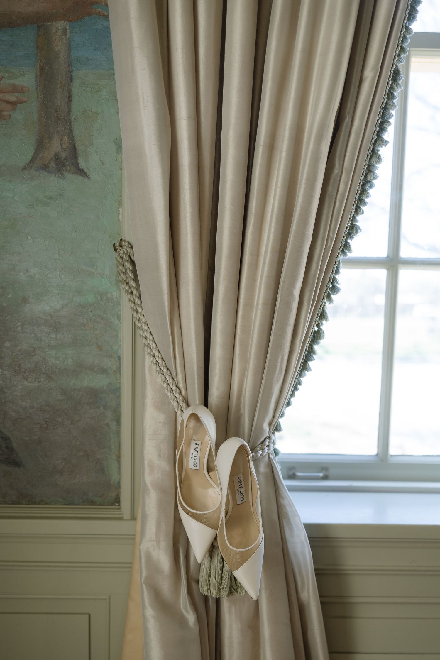 White bridal heels hang delicately from a silk curtain tieback beside a sunlit window.