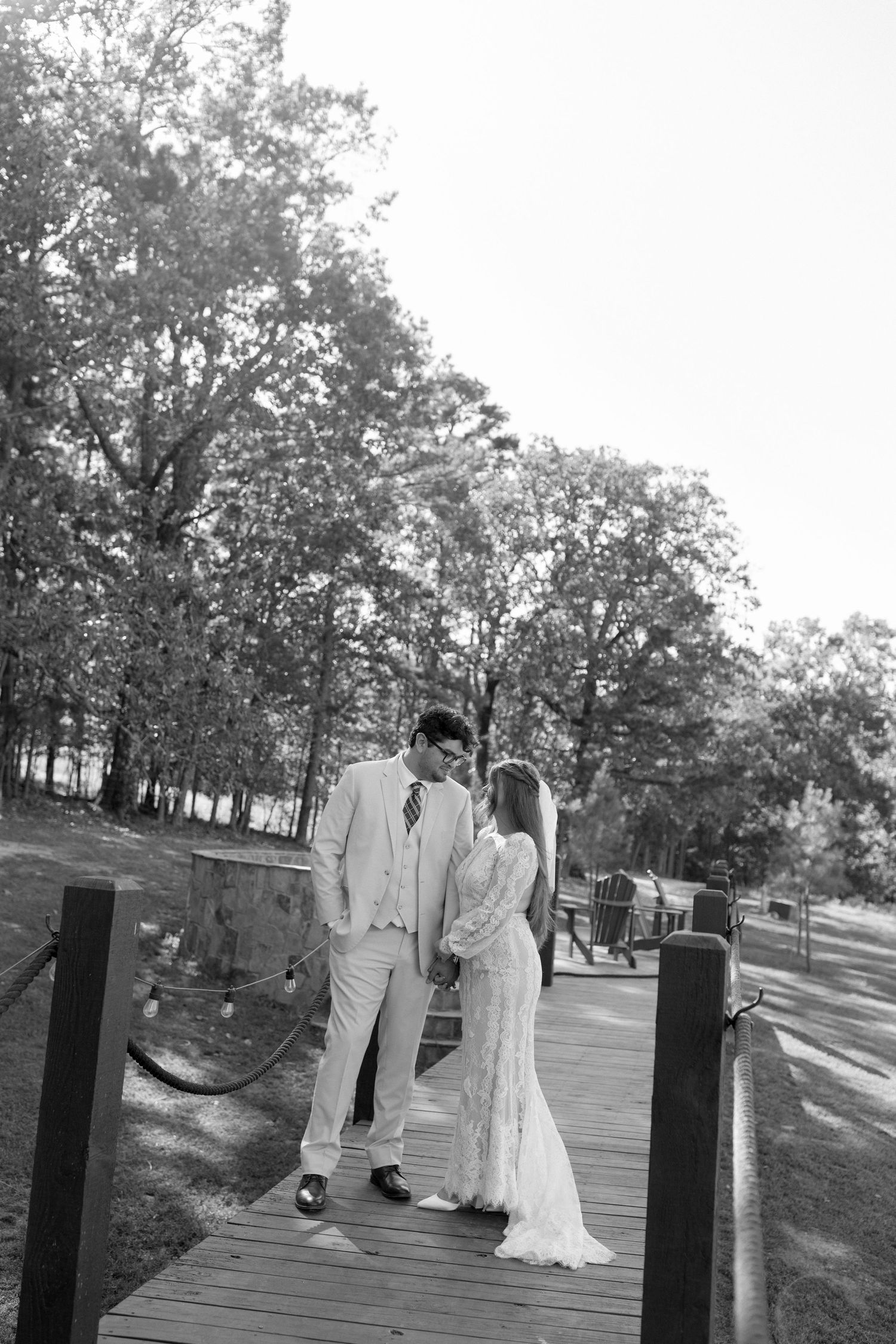 A black and white photo of a wedding couple standing on a wooden bridge surrounded by trees.