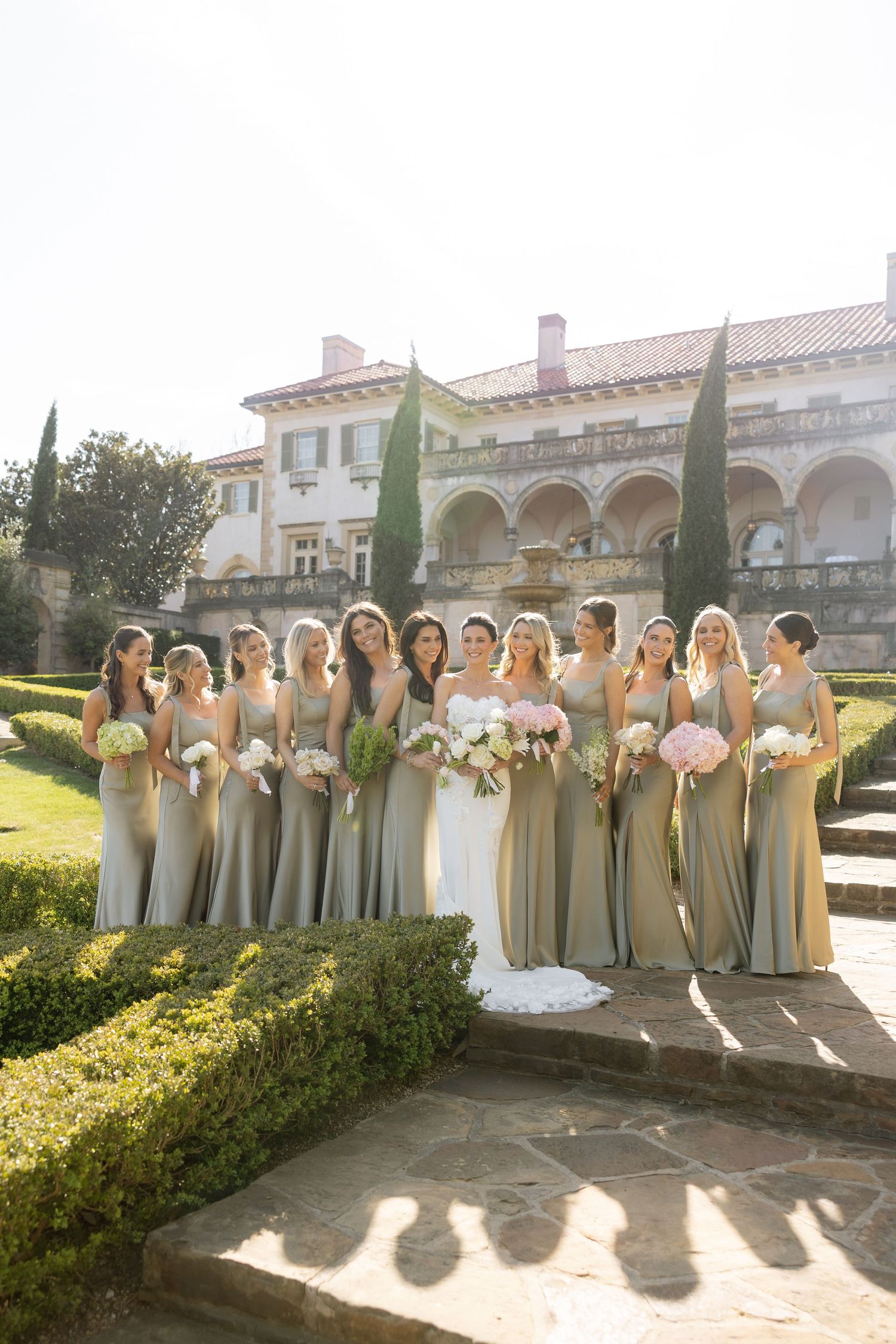 Bride poses with bridesmaids in sage green gowns holding pink bouquets outside elegant estate venue.