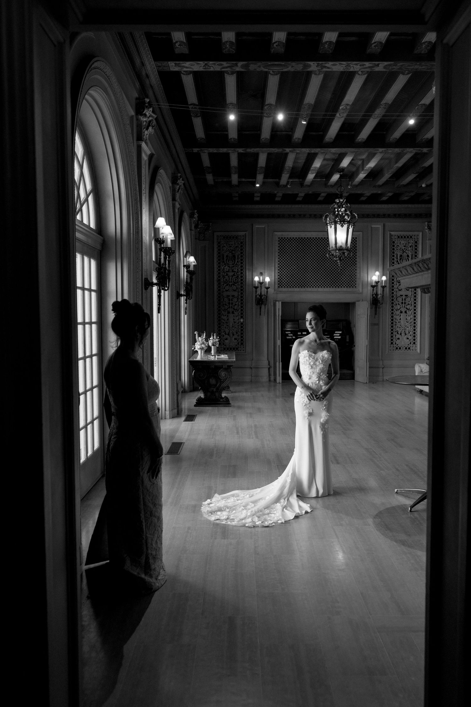 Elegant bride in fitted lace gown stands in grand ballroom hallway in black and white.