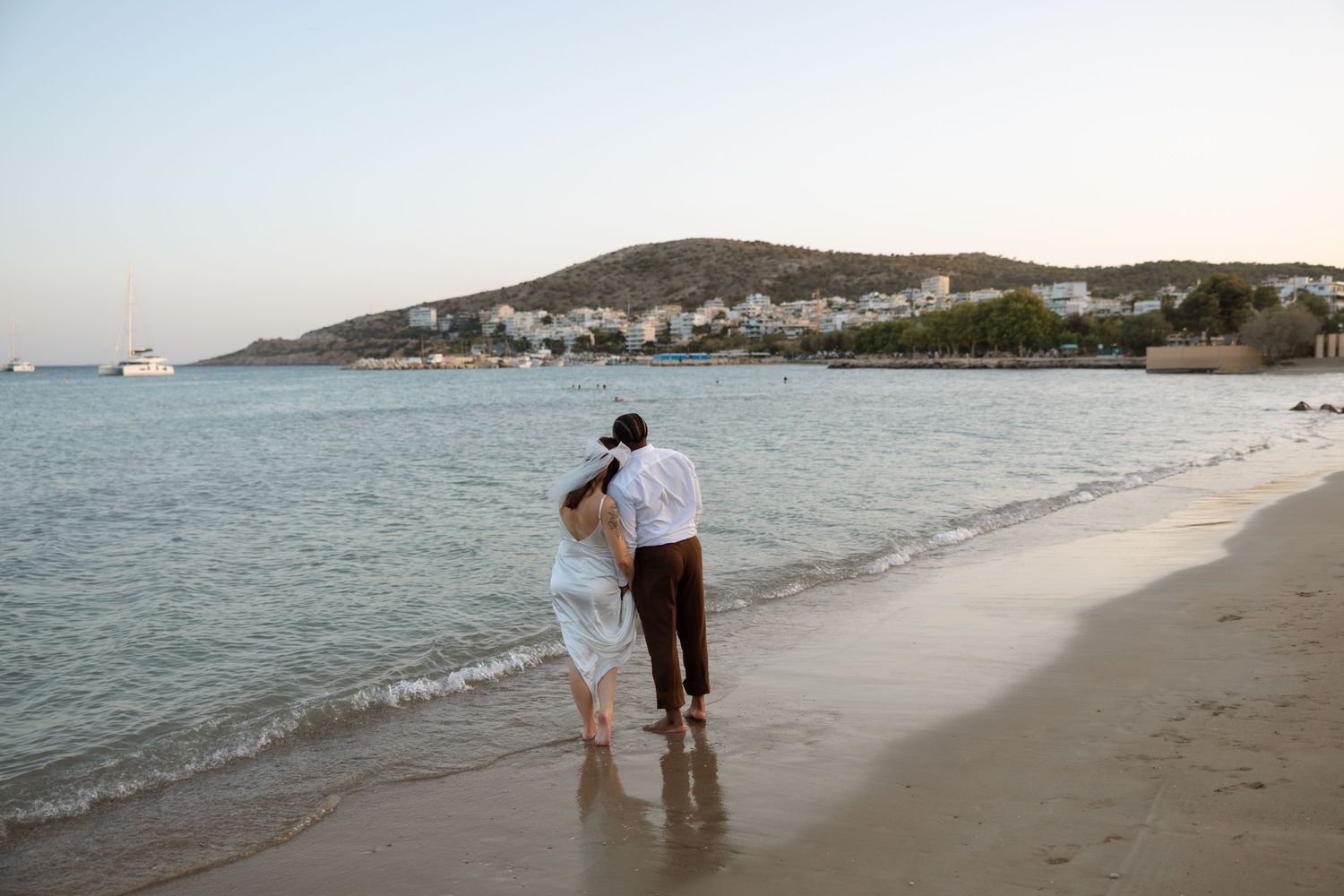 Couple stands together at water's edge on beach with hillside coastal town in background.