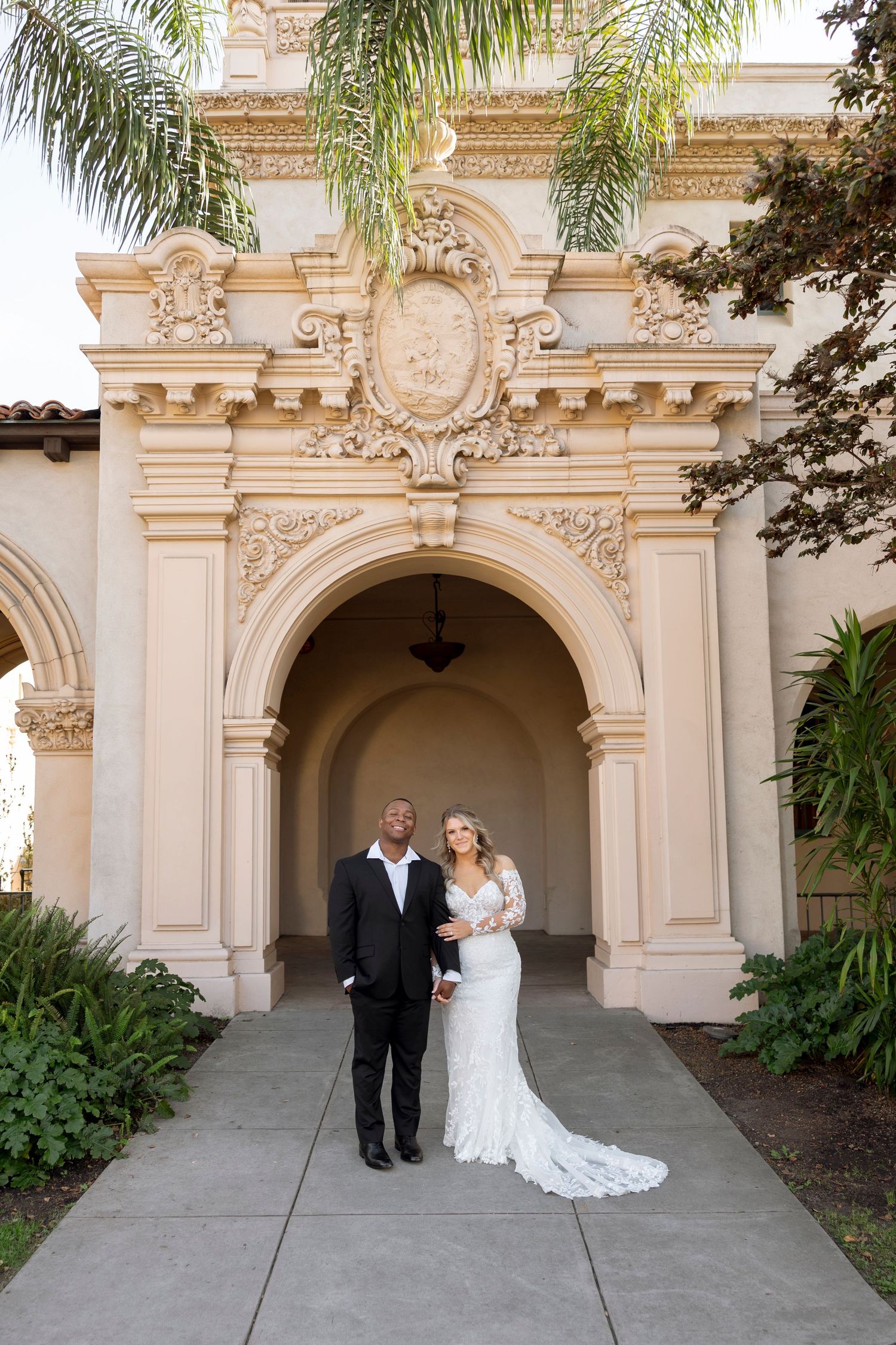 A romantic moment captured under ornate stone archways with palm trees and elegant architecture.