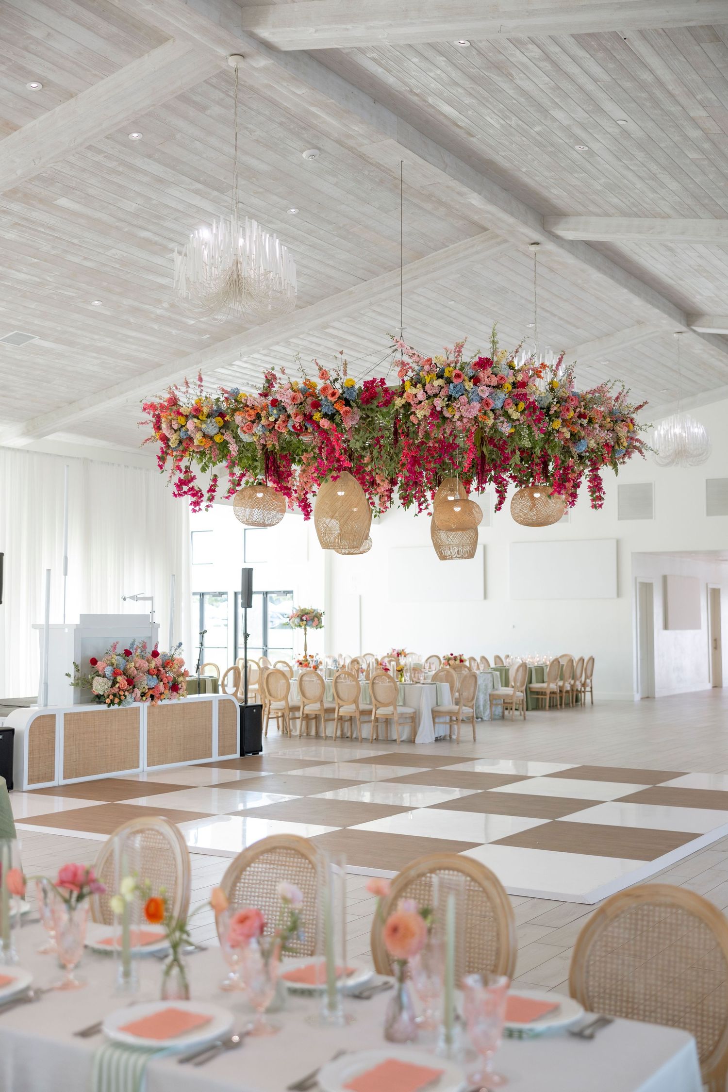 Bright wedding reception hall with hanging floral installations and cane back chairs.