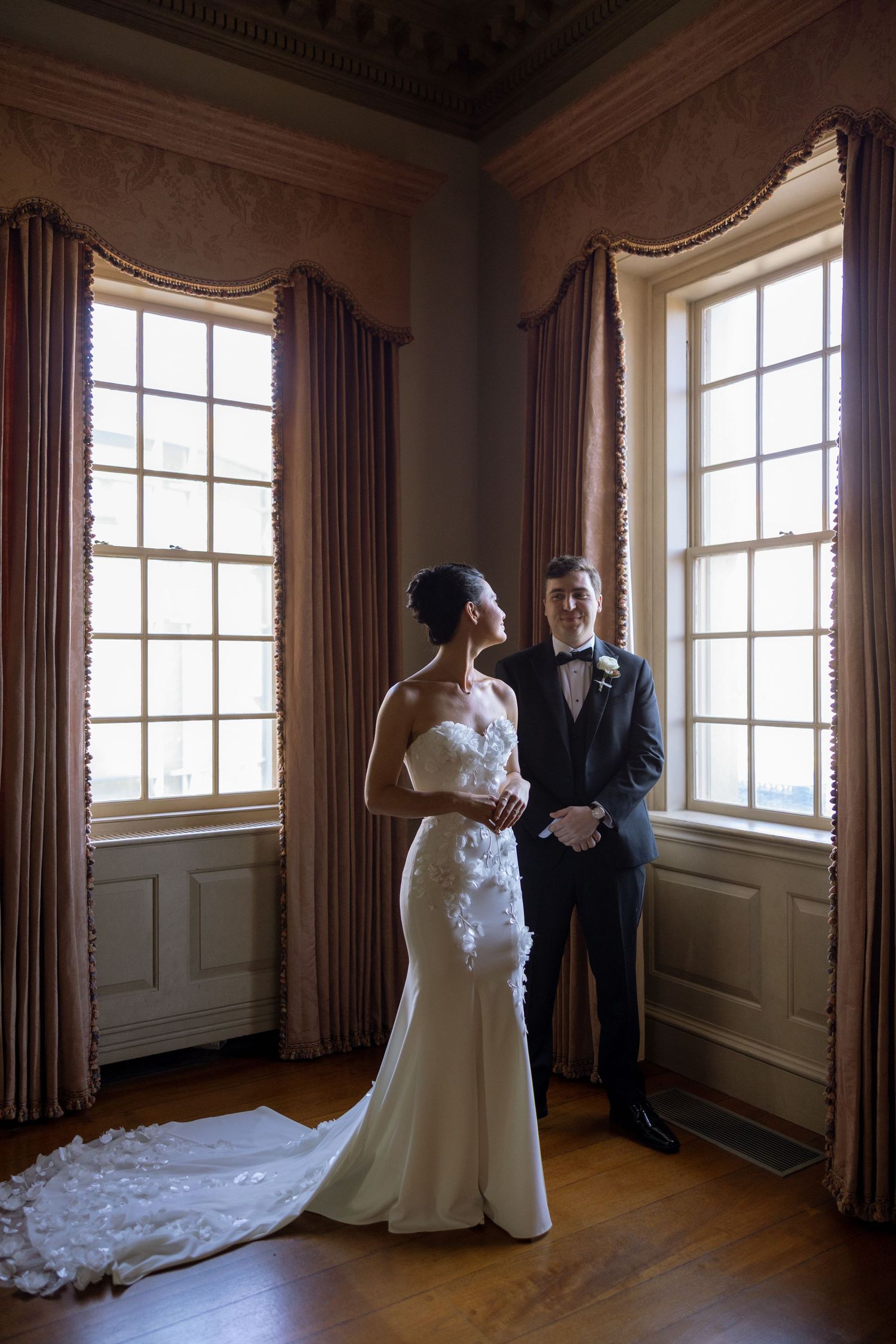 Bride in floral applique gown and groom pose by tall windows in elegant historic estate room.