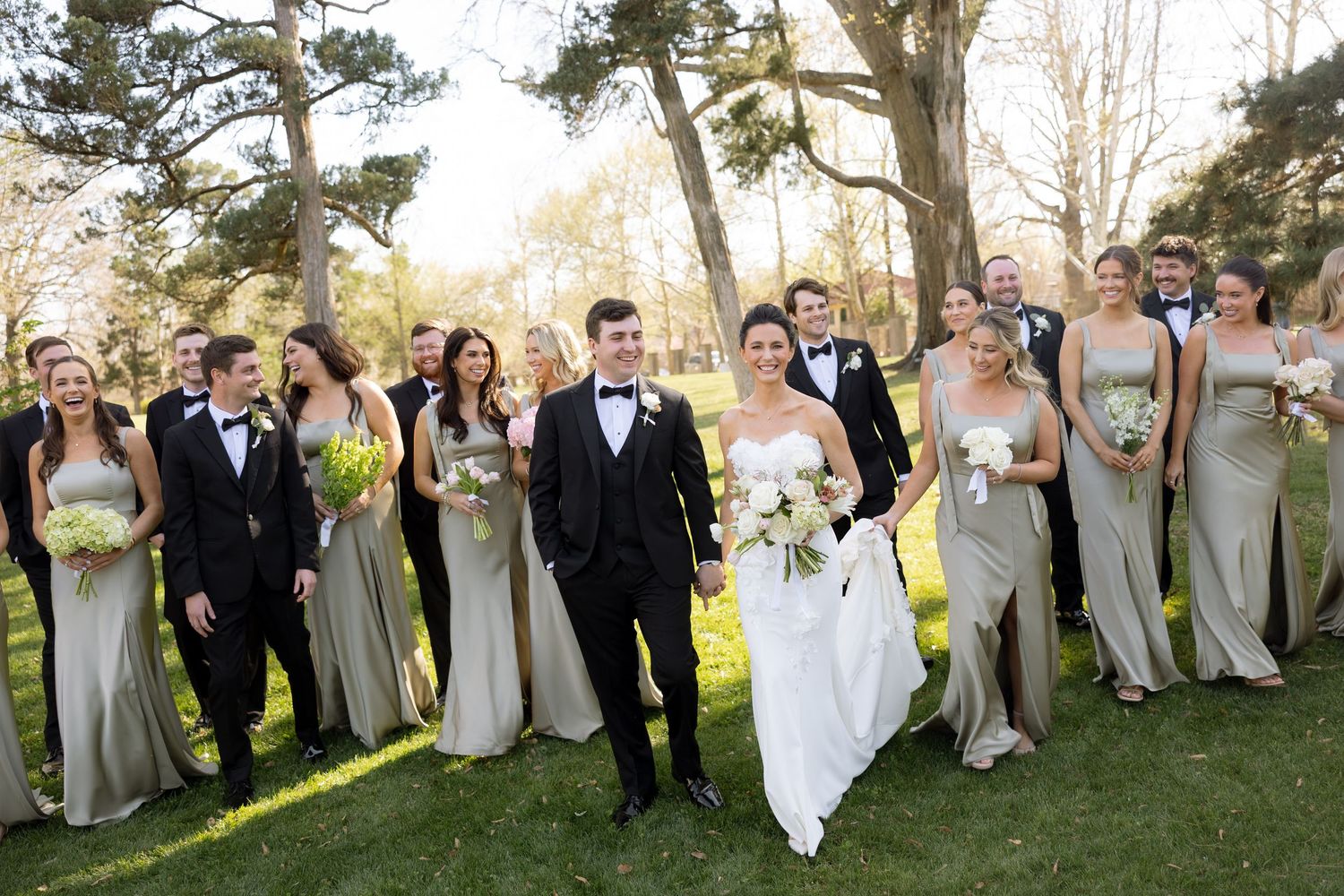 Large wedding party poses outdoors, bridesmaids in champagne gowns and groomsmen in black tuxedos.