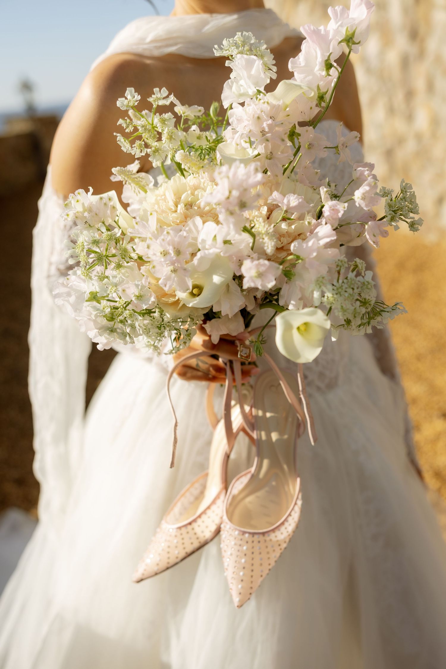 Bride holding a white floral bouquet with blush ribbons and matching pointed-toe heels in a white wedding gown.