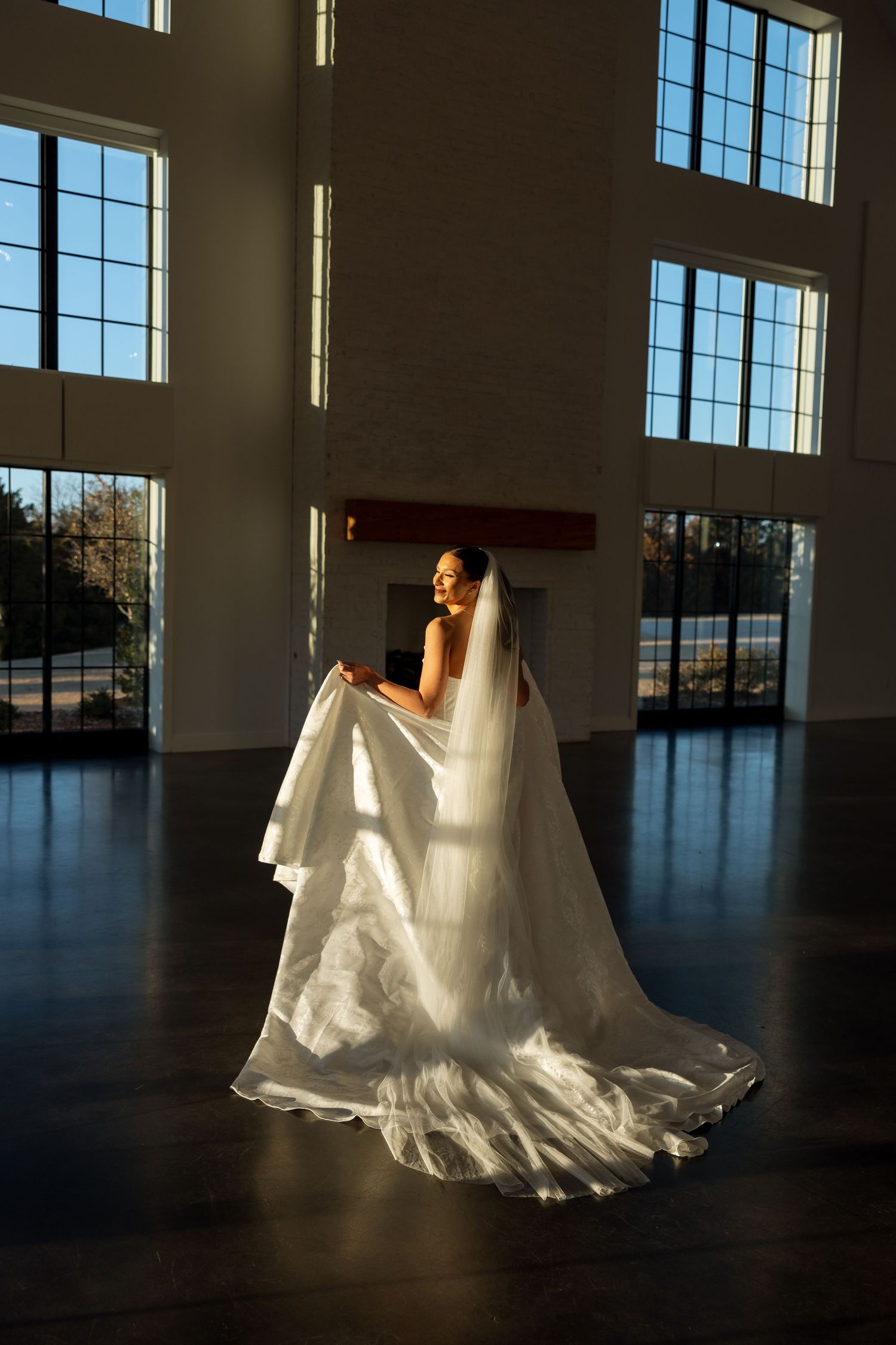 A bride in a white wedding gown stands dramatically in sunlight streaming through tall windows.