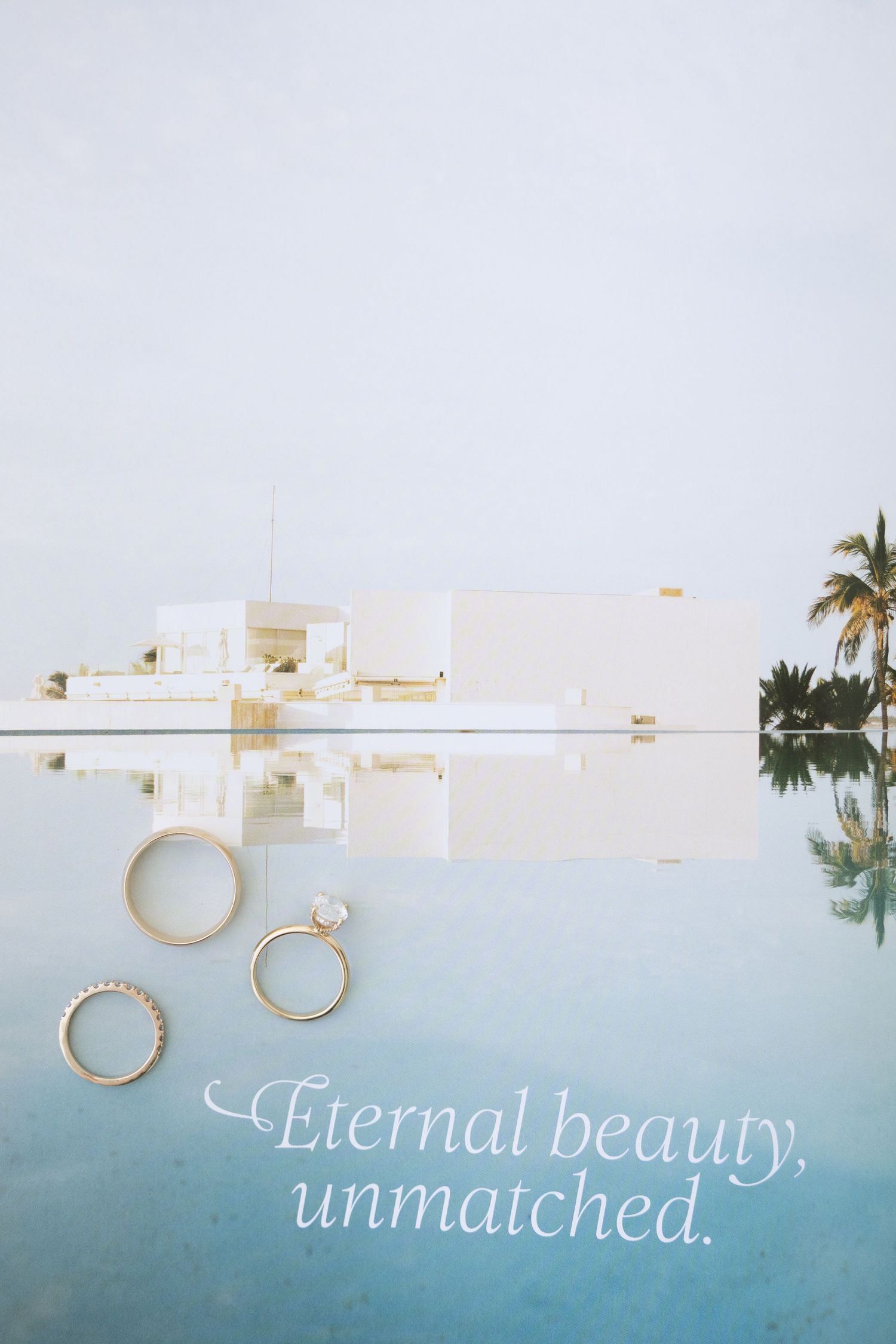 Gold wedding rings displayed near infinity pool with white building reflection and palm trees.