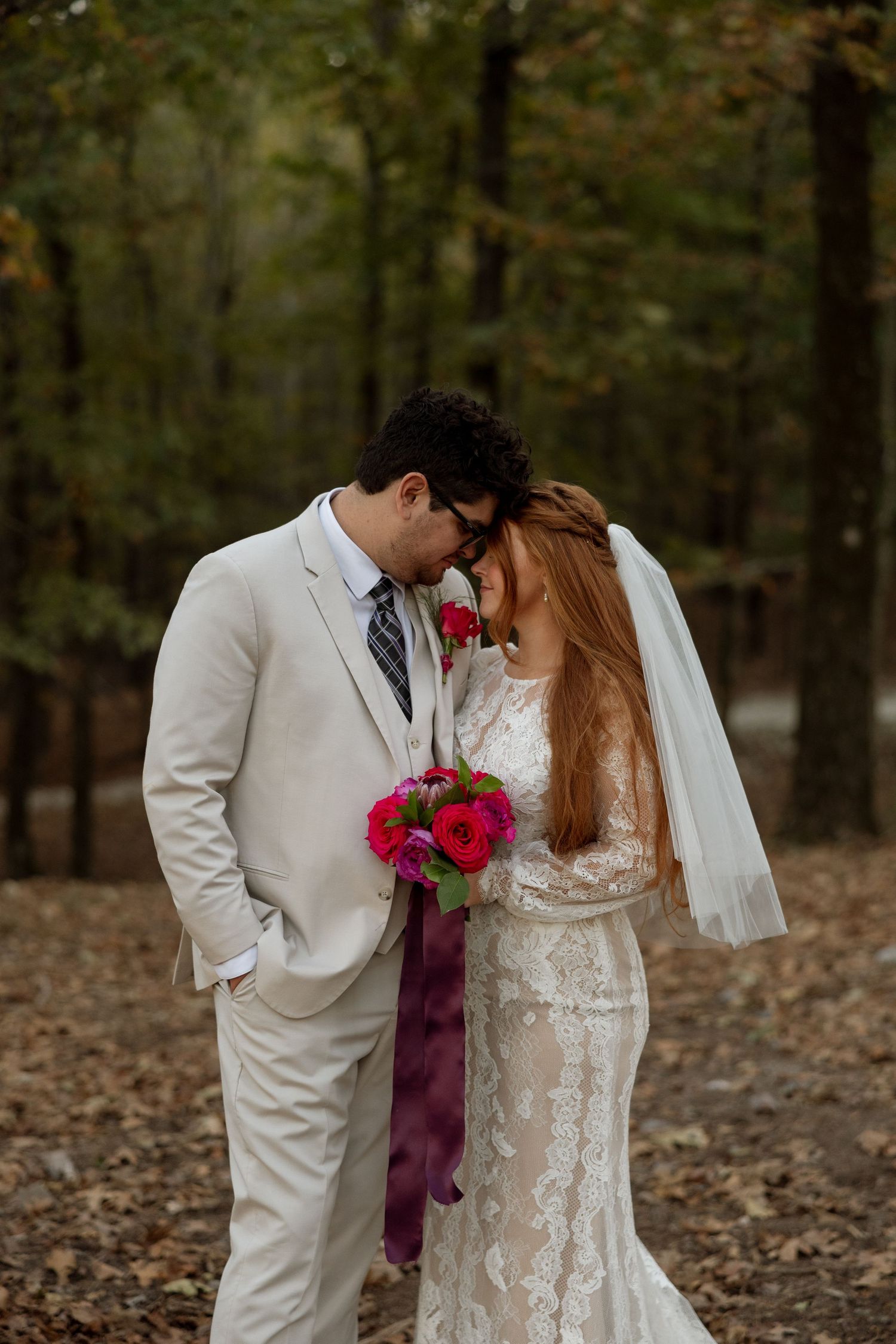 A romantic wedding couple embraces in an autumn forest setting while holding a vibrant pink and red bouquet.