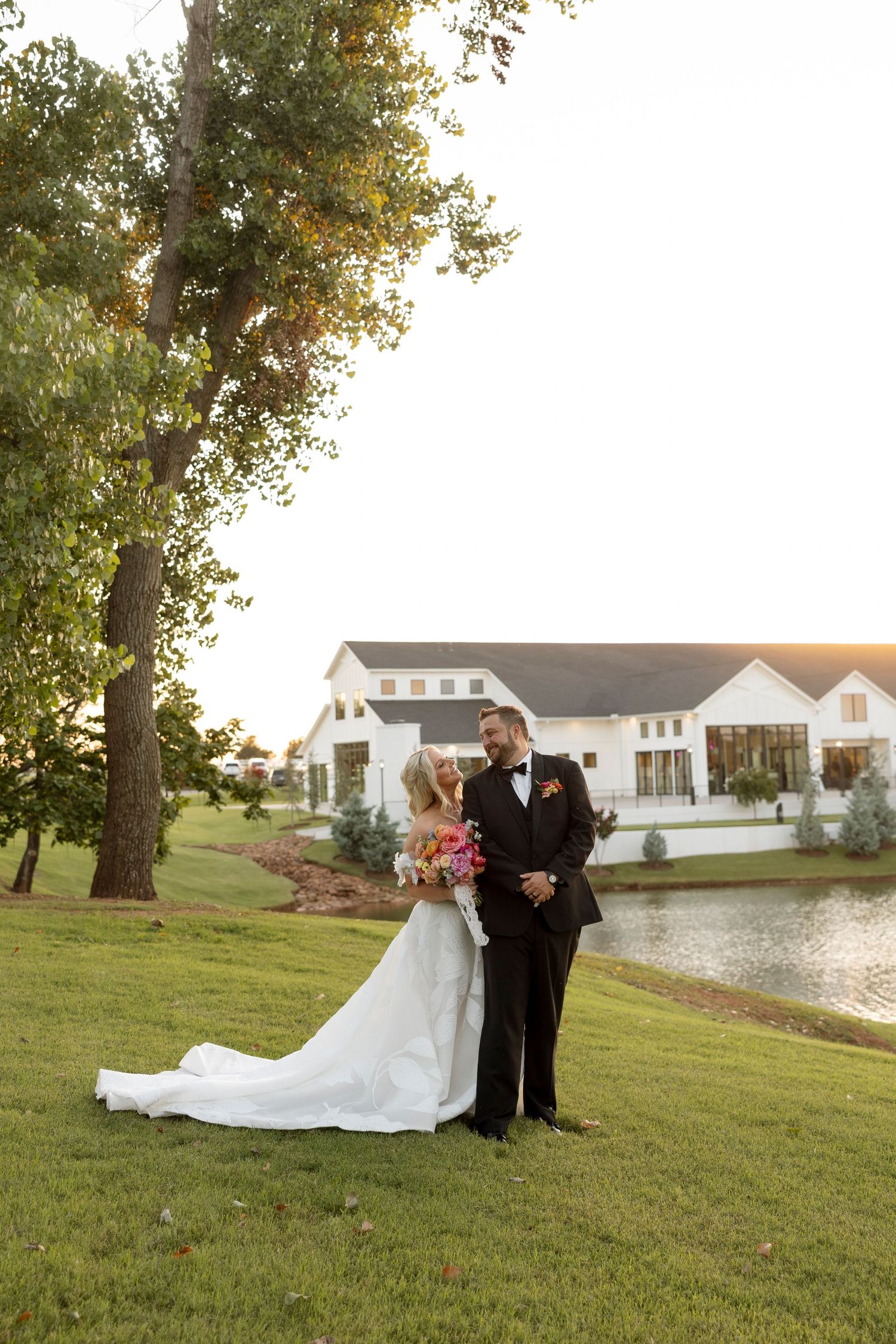 Wedding couple poses on lawn in front of white farmhouse venue with pond at sunset.