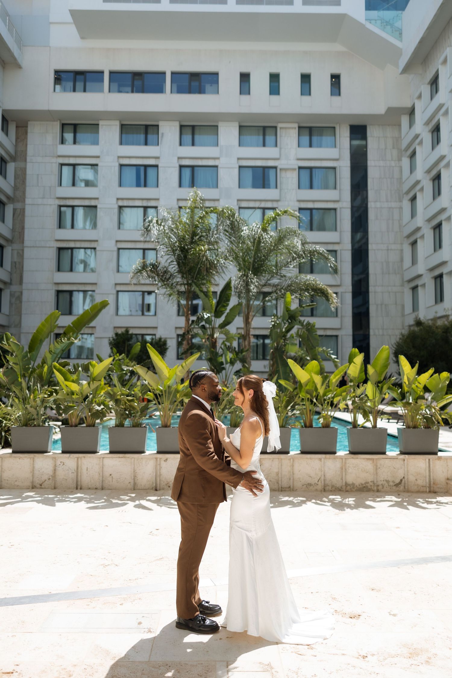 Newlyweds embrace in front of a luxury hotel with tropical landscaping.