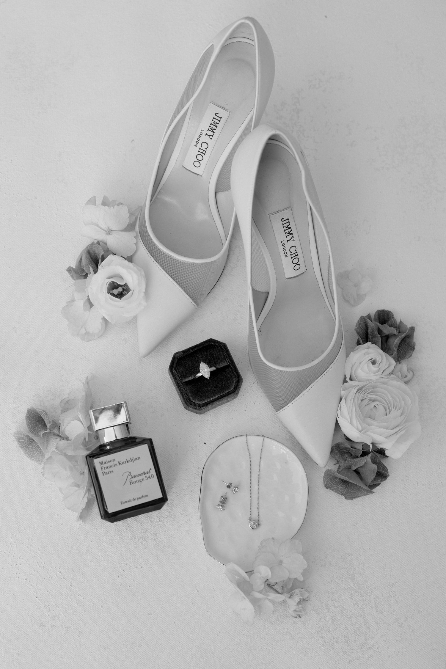 Black and white flat lay of Jimmy Choo bridal heels, engagement ring, perfume, and floral accents.