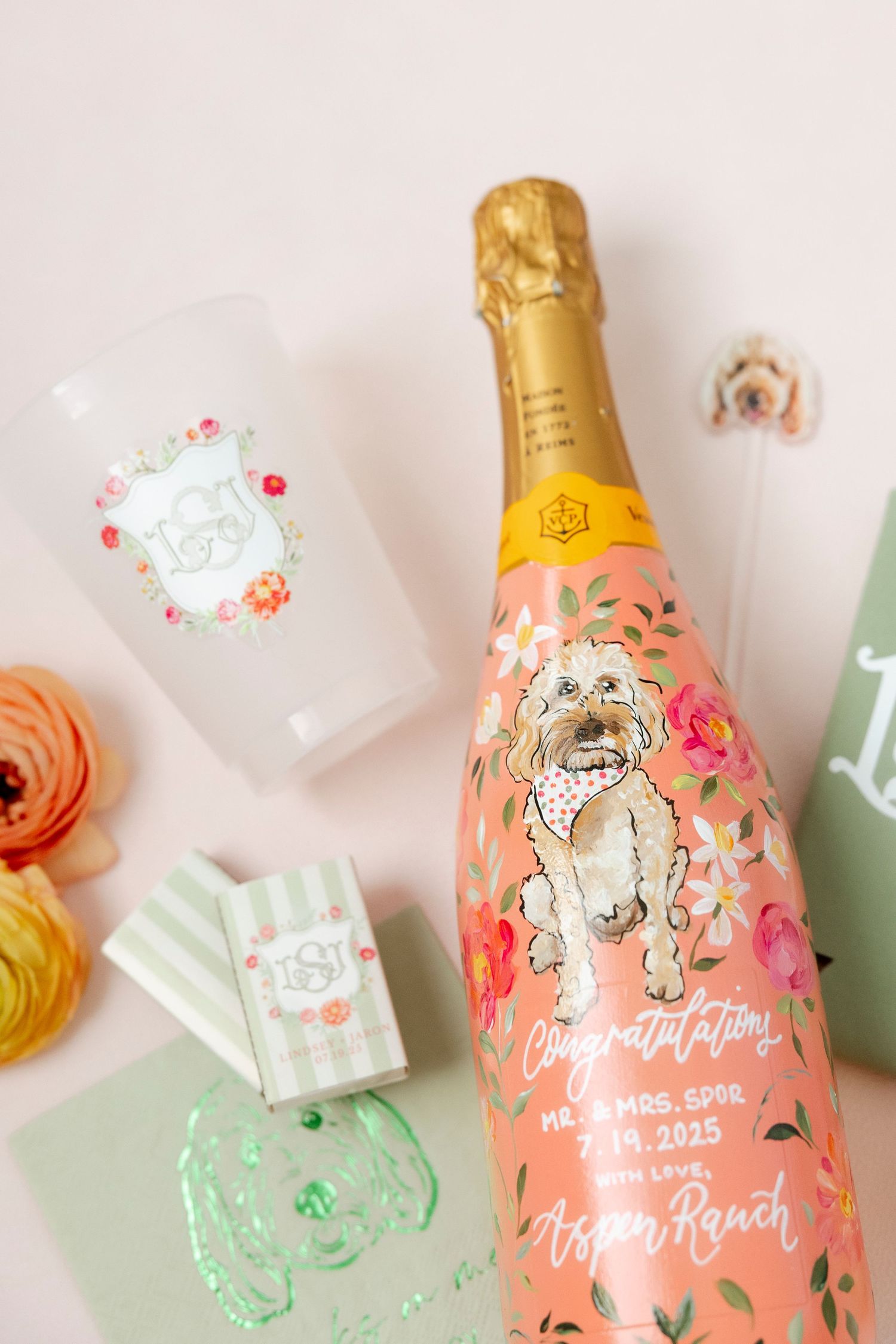 Custom champagne bottle with illustrated dog design and floral decorations alongside matching party cups and ranunculus flowers.