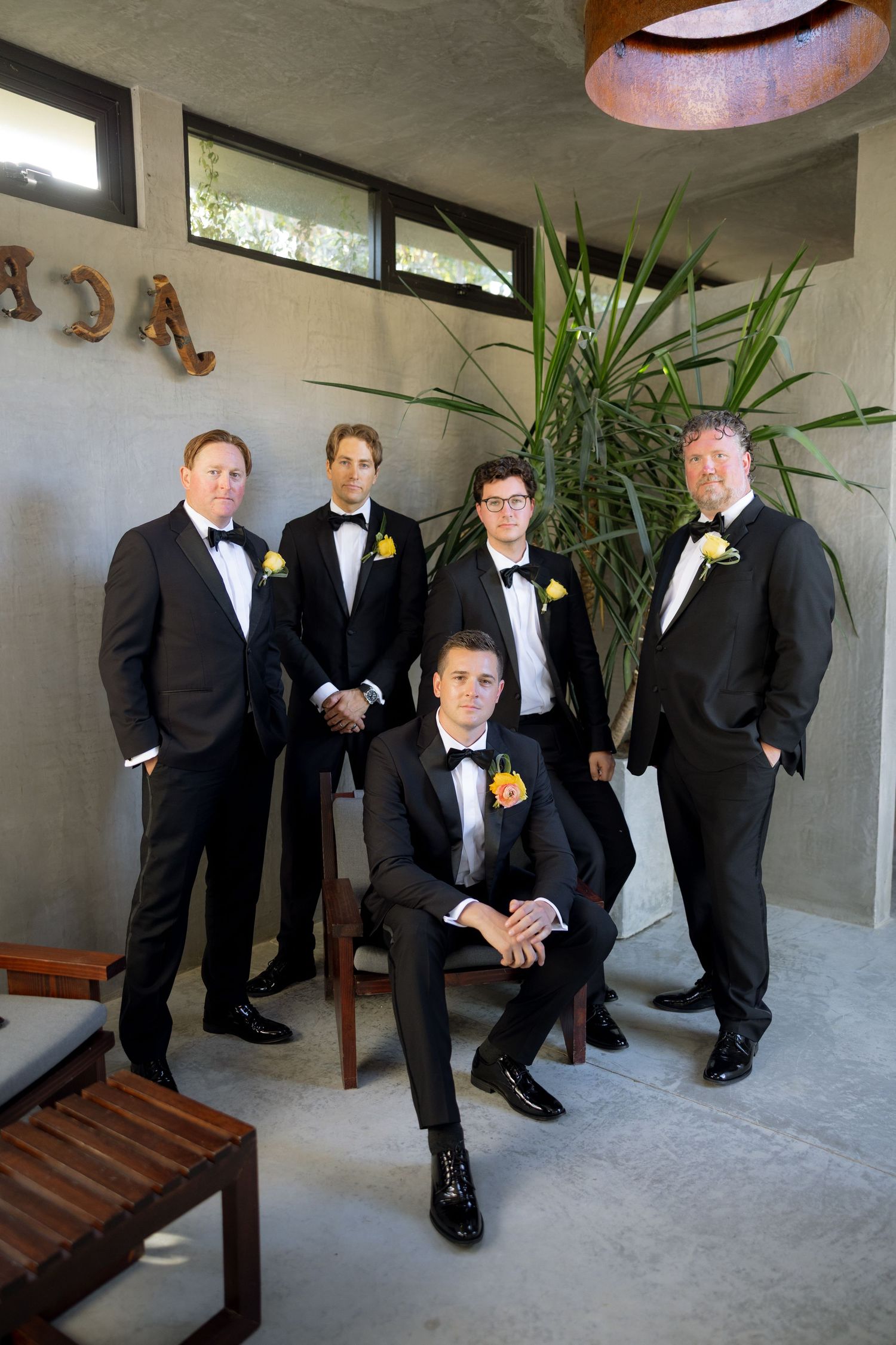 Five groomsmen in black tuxedos with yellow boutonnieres pose for a formal wedding photo indoors.