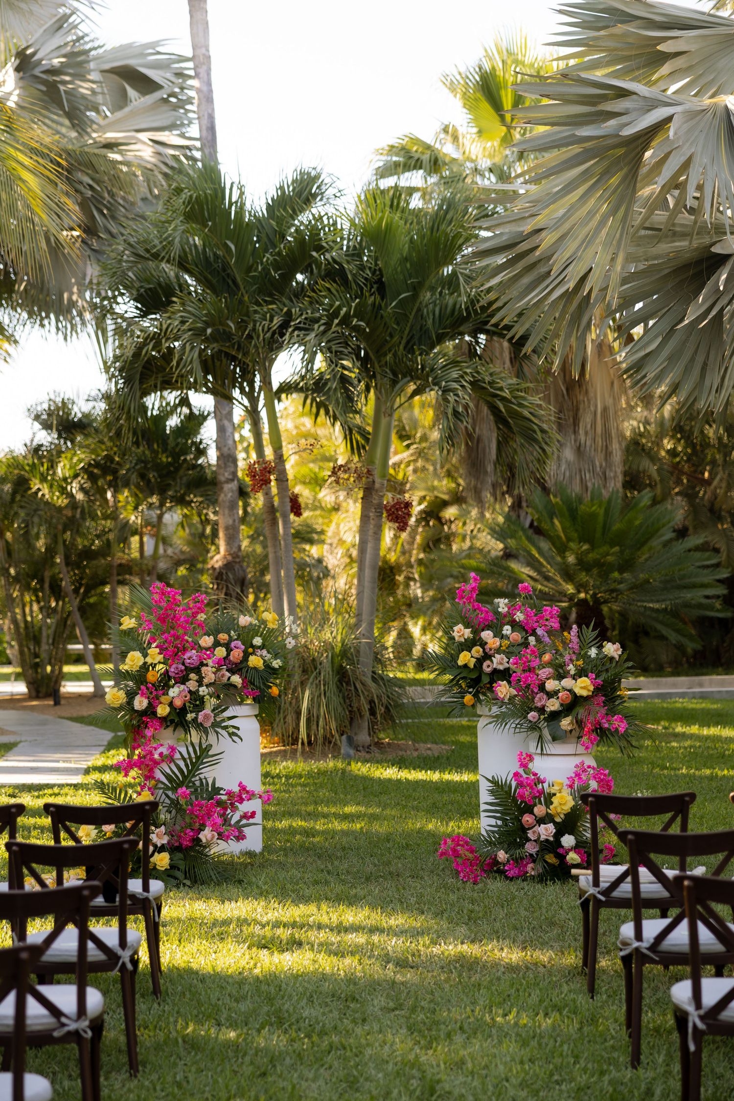 Vibrant pink and yellow floral ceremony arch with wooden chairs on green lawn amid palm trees.