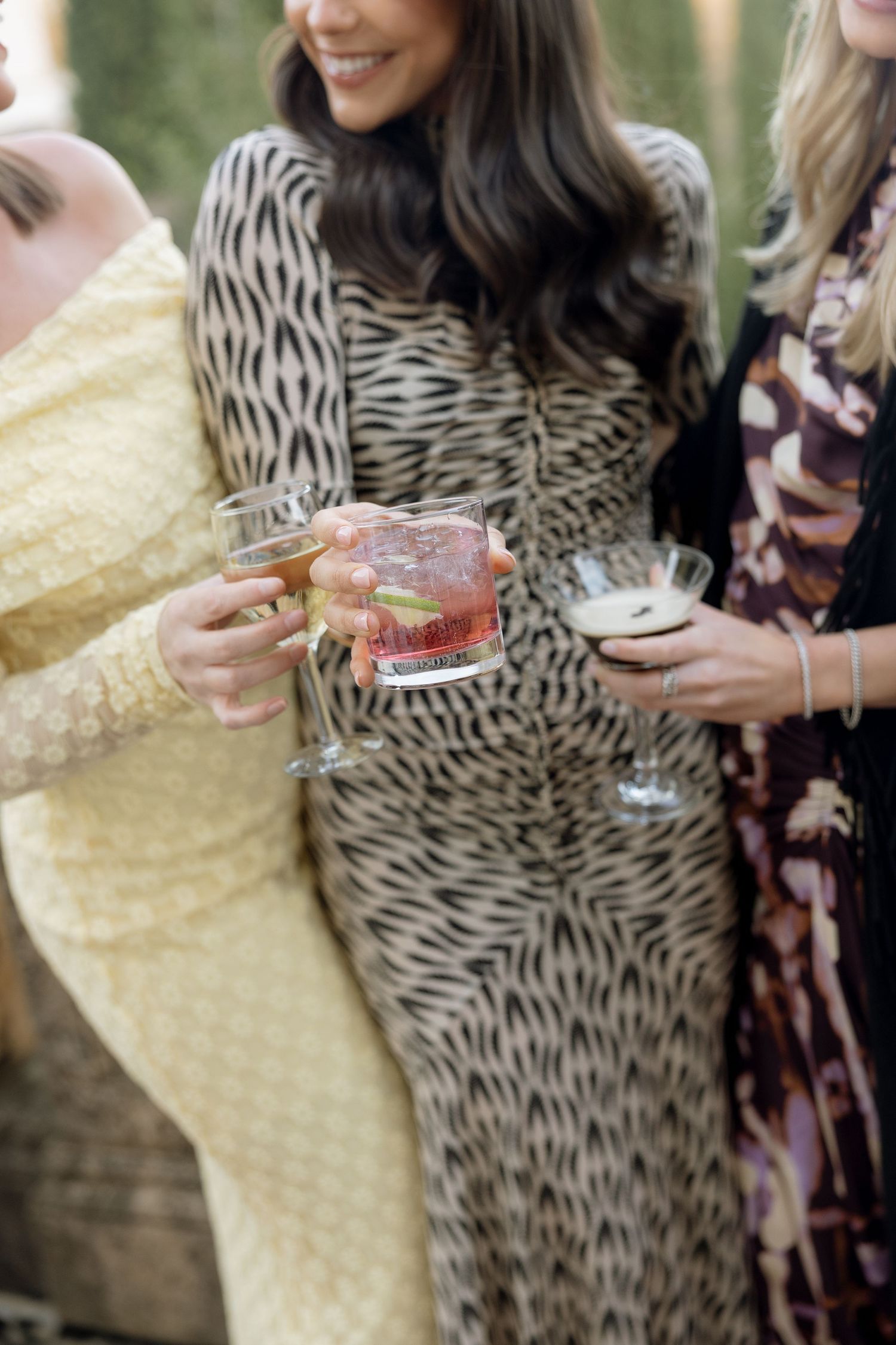 Wedding guests in elegant dresses clink colorful cocktail glasses together at an outdoor celebration.