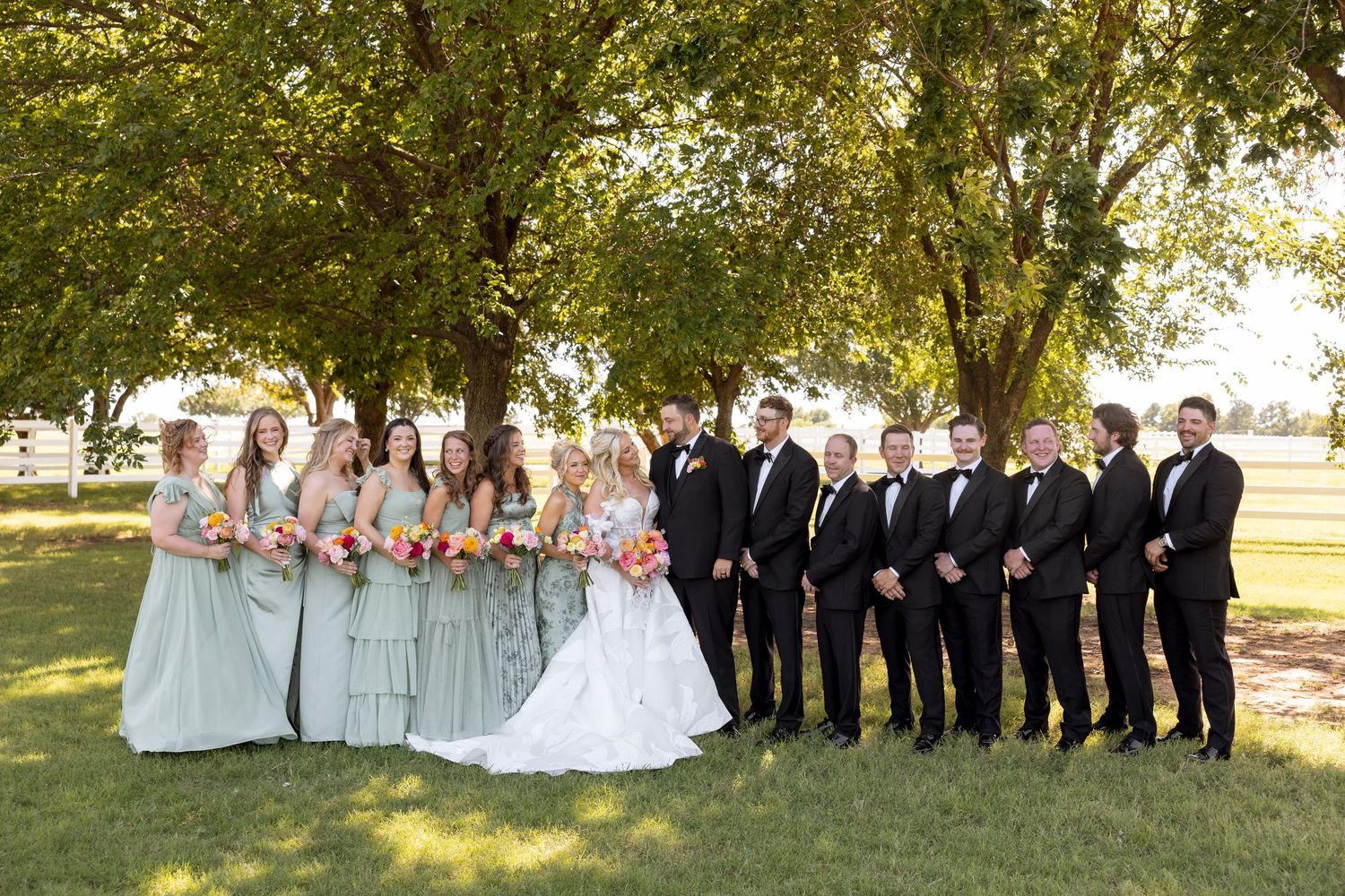 Wedding party poses outdoors under green trees in formal attire.