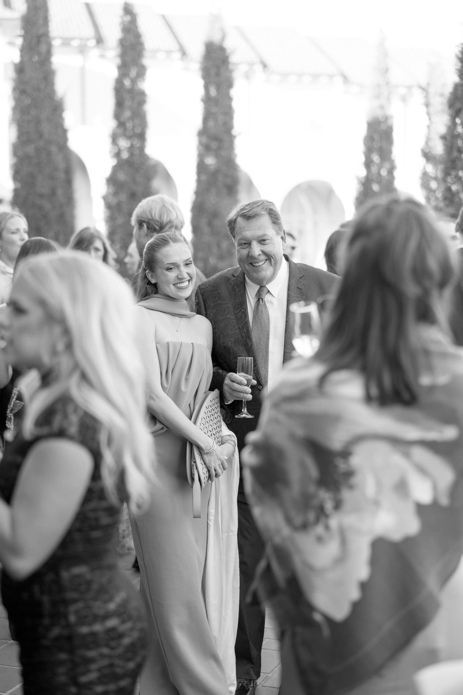 Black and white candid of smiling wedding guests socializing outdoors near tall cypress trees.