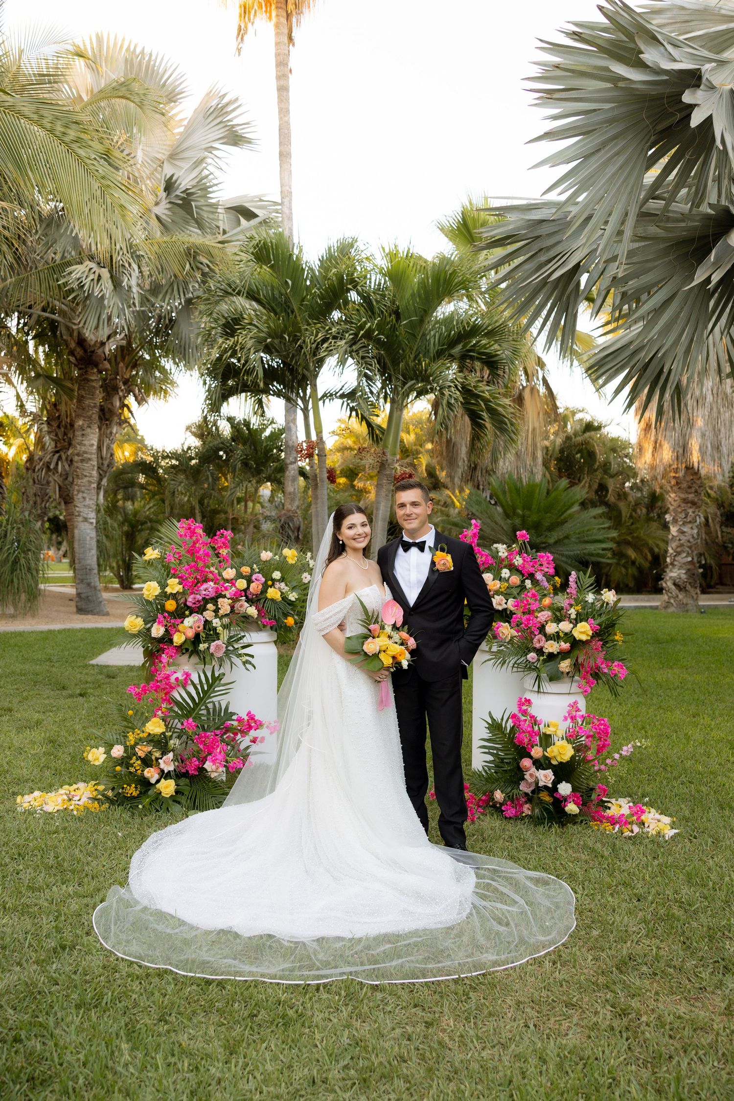 Bride and groom pose before vibrant pink and yellow floral arch surrounded by tropical palm trees.