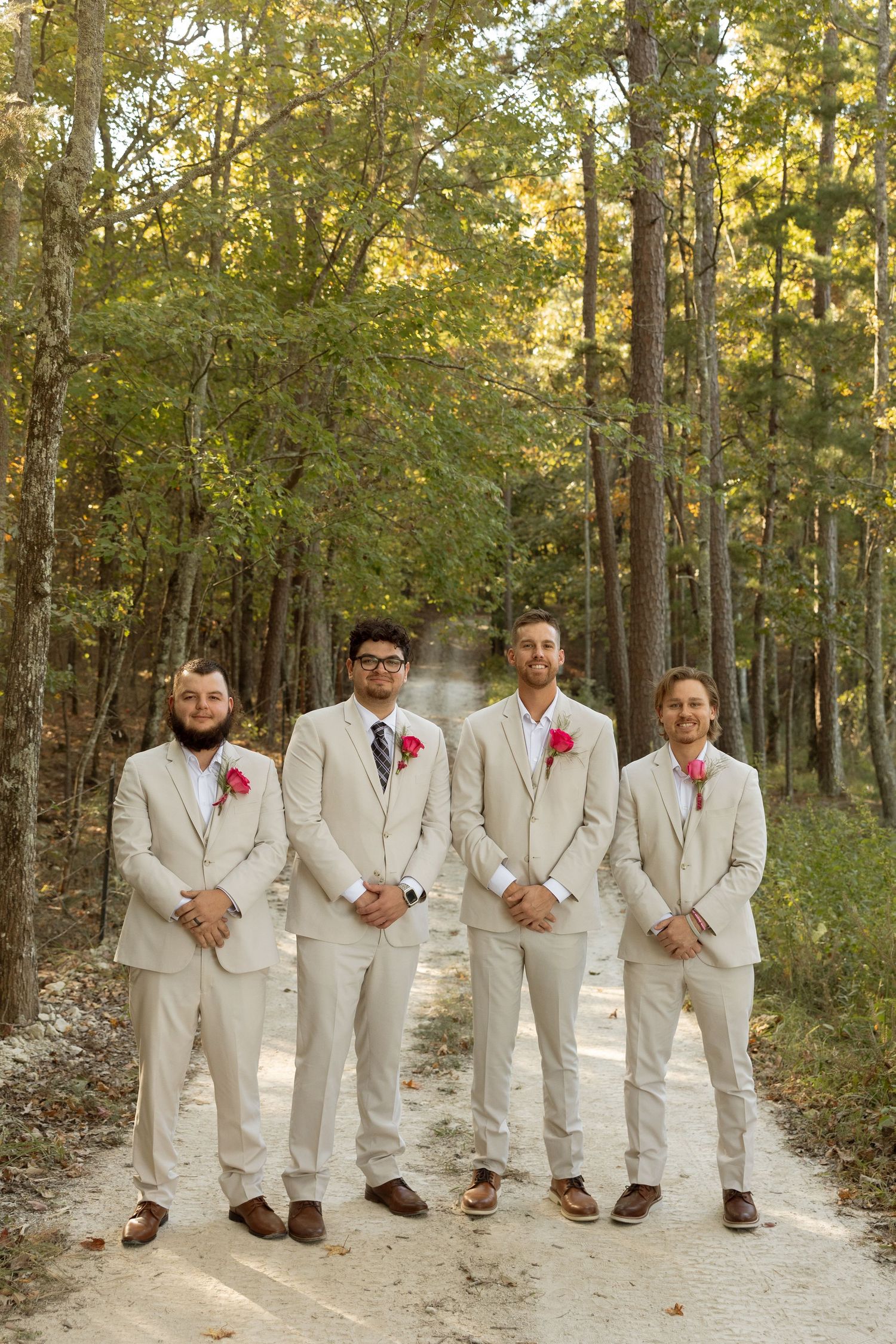Four groomsmen in cream-colored suits with pink boutonnieres pose on a forest path.