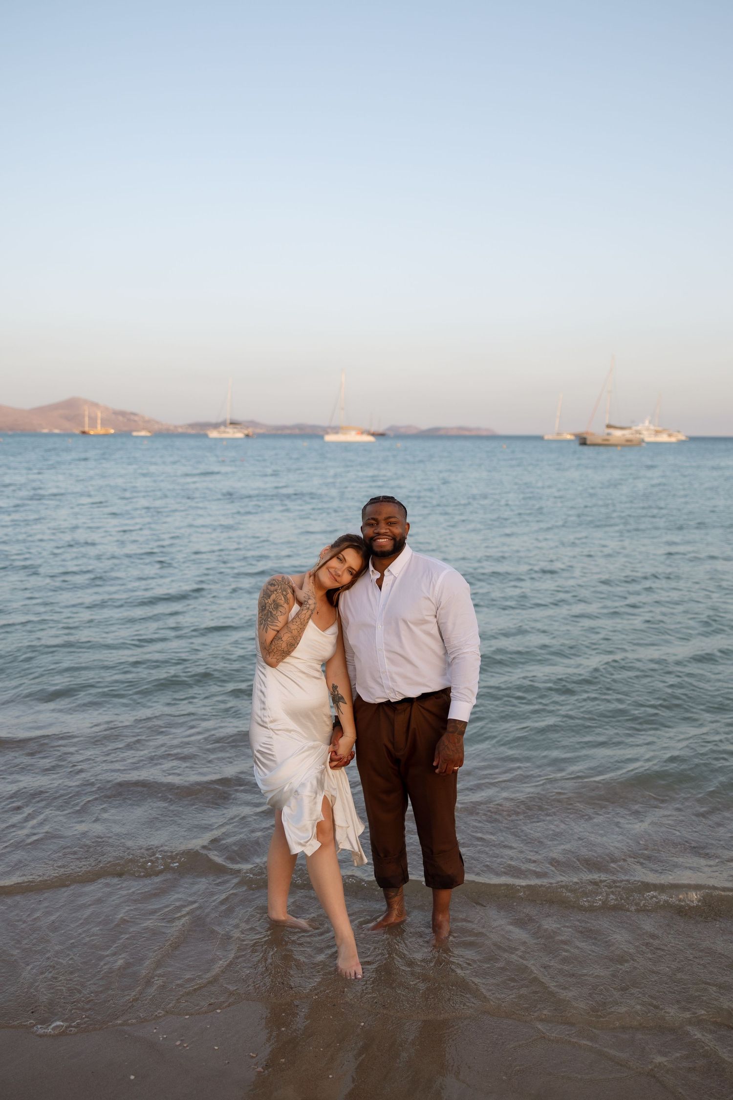 A couple stands together in shallow water with boats anchored in the tranquil bay behind them.
