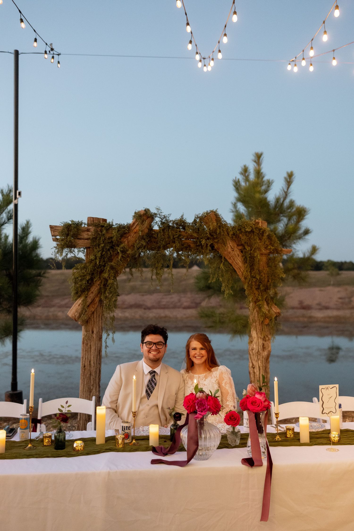 Newlyweds stand at an elegantly decorated outdoor reception table with string lights and floral arch at sunset by a lake.