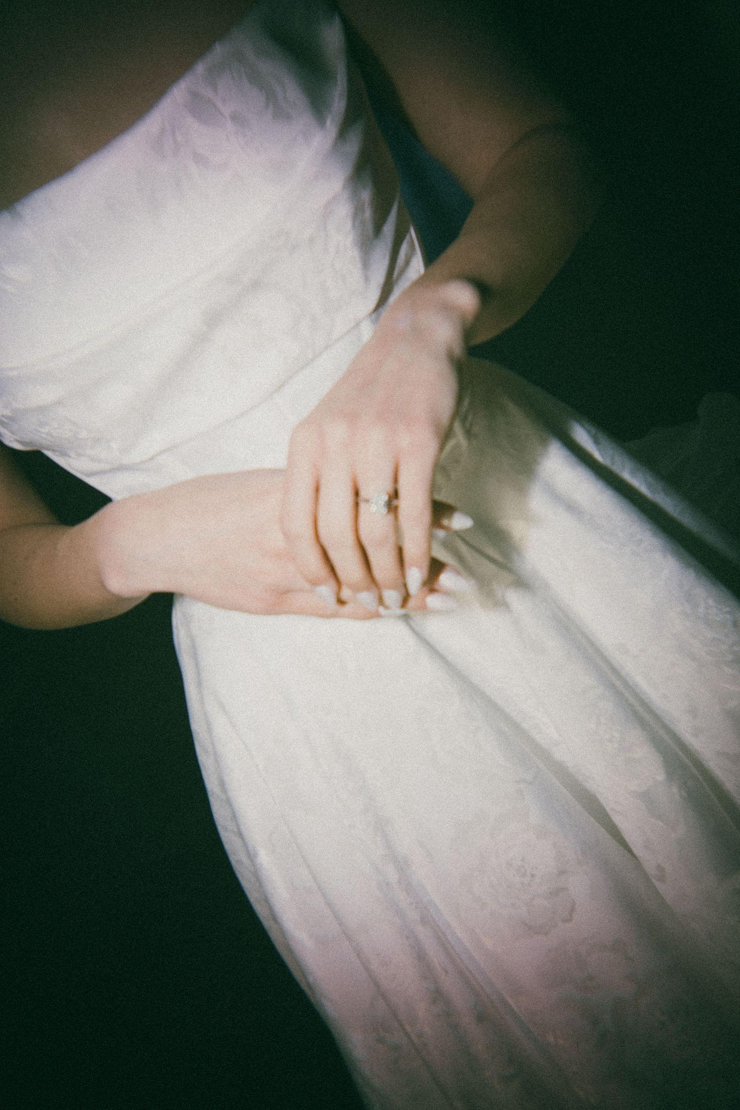 A delicate hand with an engagement ring rests on a flowing white wedding dress in a dreamy, vintage-style photograph.