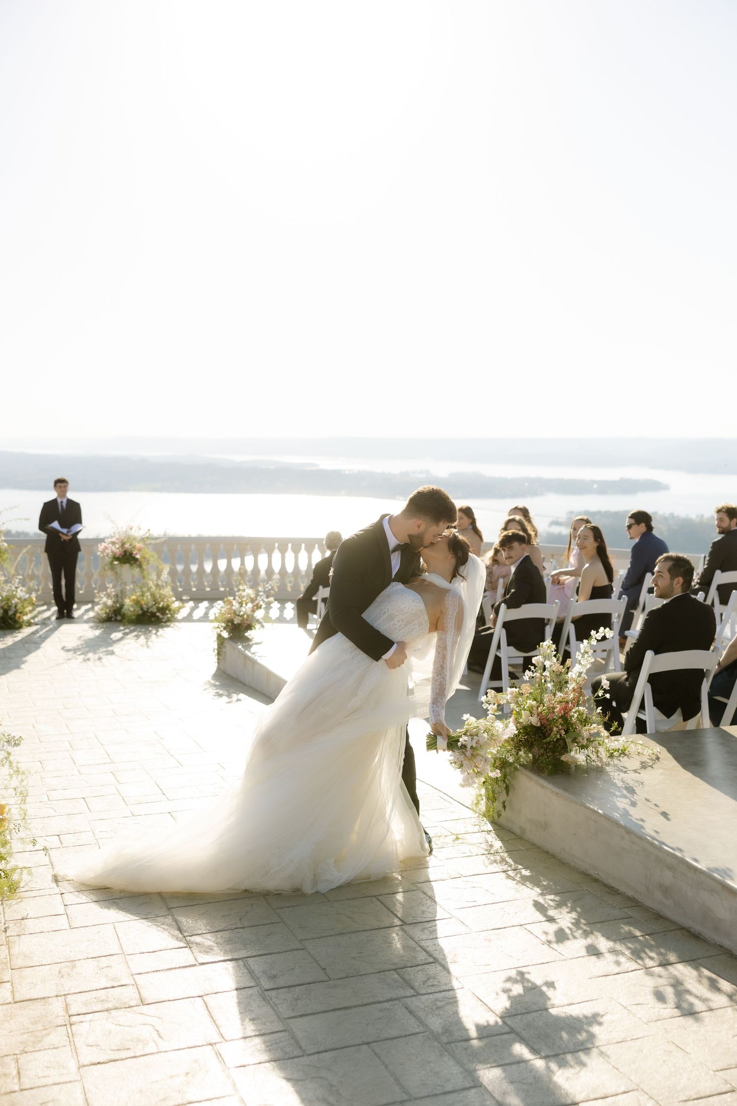 Bride and groom share first kiss dip at outdoor wedding ceremony with scenic waterfront views and seated guests.