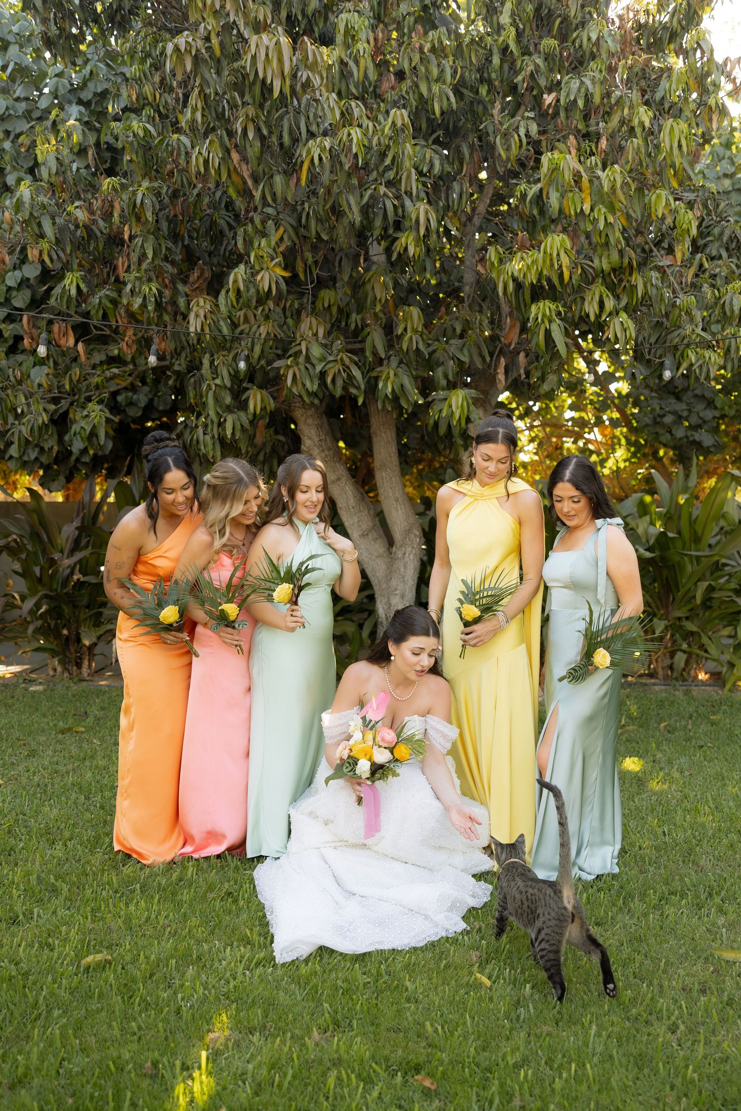 Bride sits on grass with bridesmaids in colorful mismatched gowns holding tropical bouquets near a large tree.