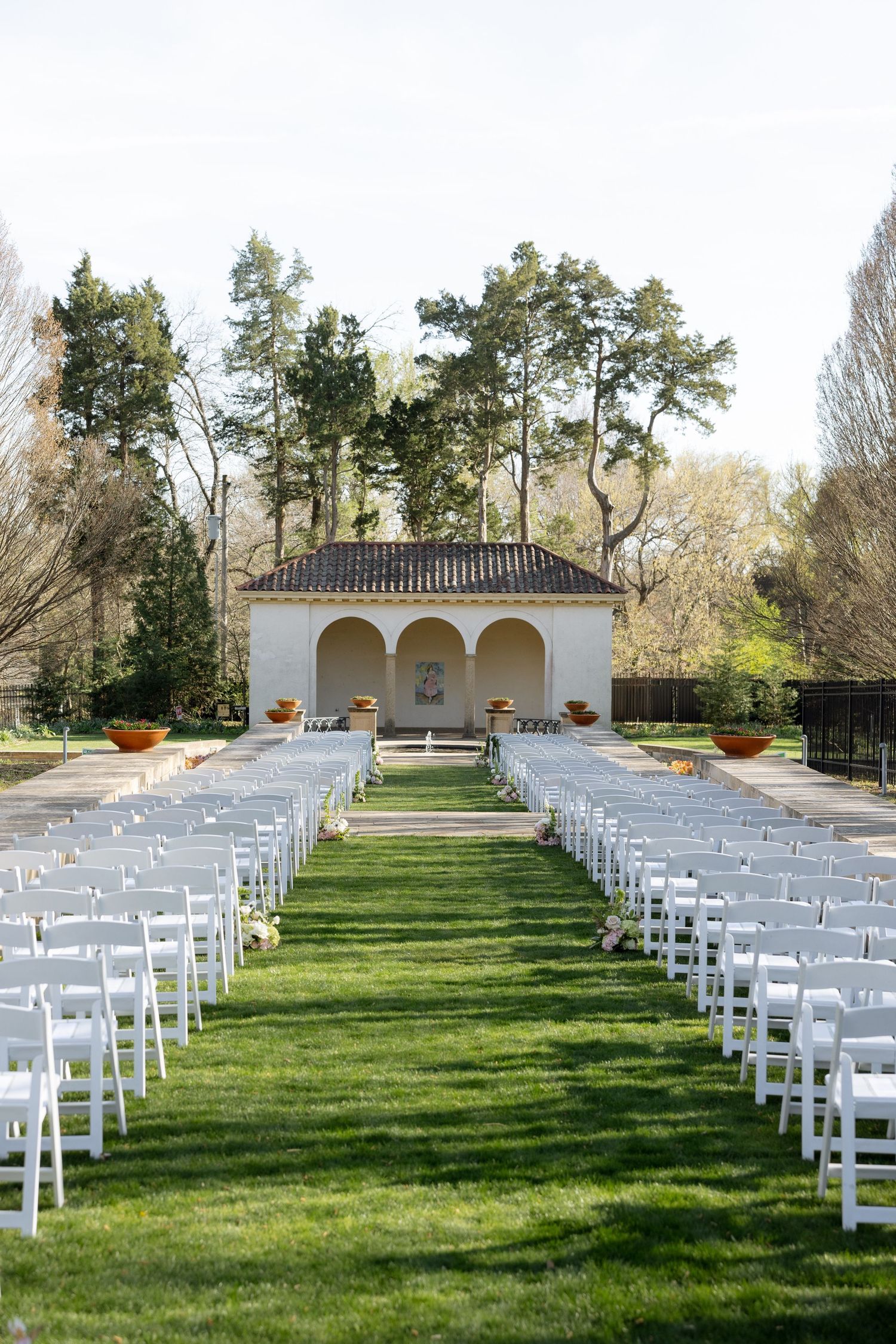 Elegant outdoor wedding ceremony setup with white chairs on green lawn leading to a white stucco pavilion surrounded by trees.